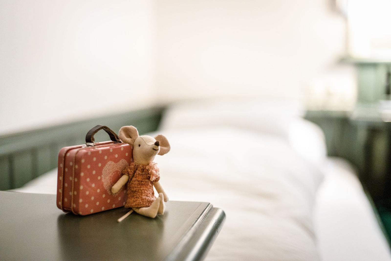 Ein Stoffmaus mit Koffer sitzt auf einem Tisch vor einem Bett.