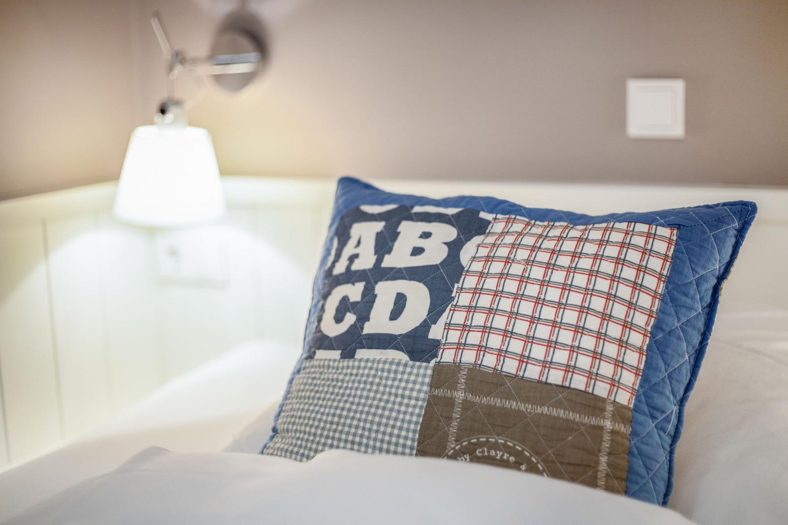 Ein Kissen mit Patchwork-Muster liegt auf einem Bett. Eine Wandlampe ist im Hintergrund sichtbar.