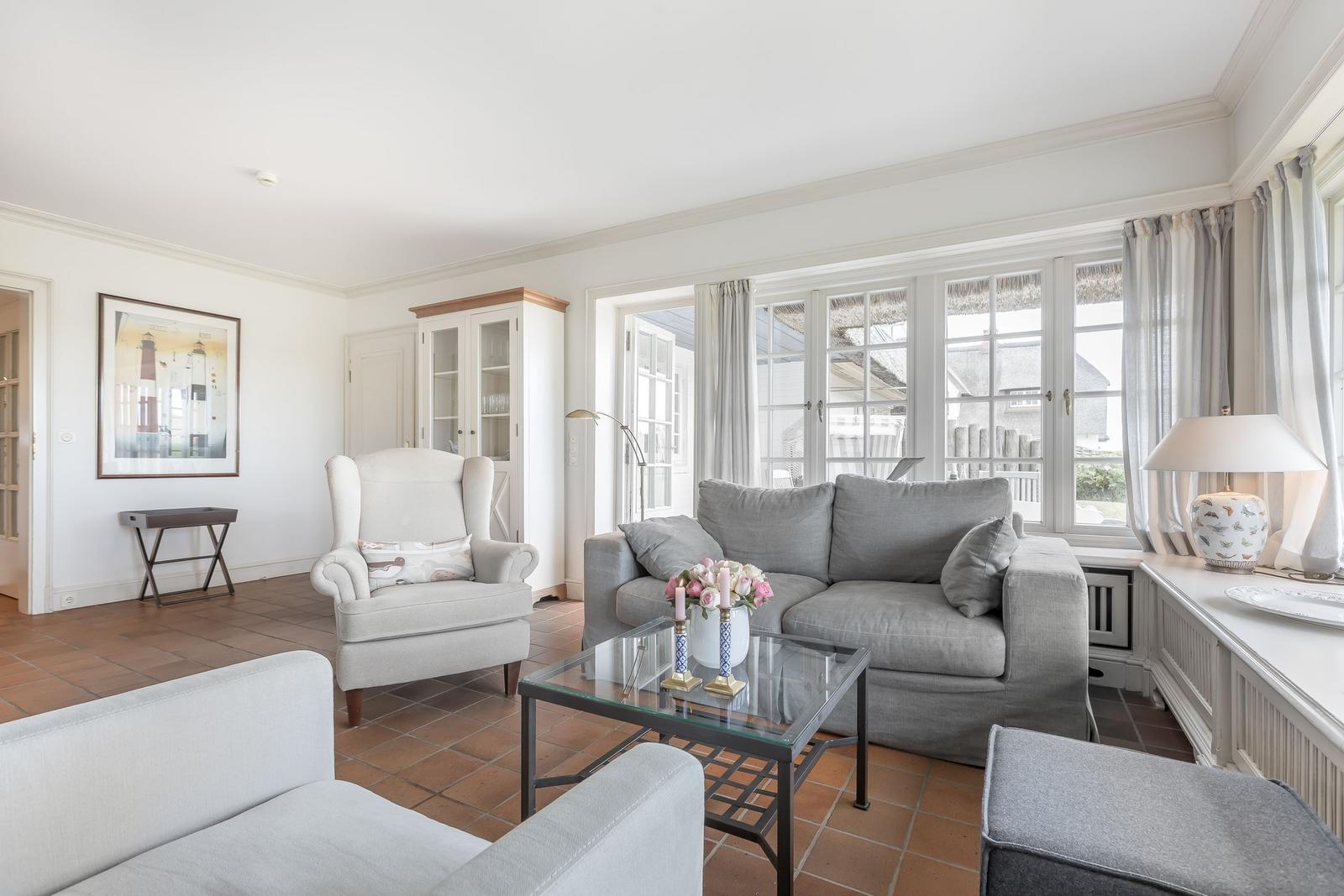 Wohnzimmer mit grauem Sofa, weißem Sessel und Glas-Tisch. Große Fenster und Terrasse.