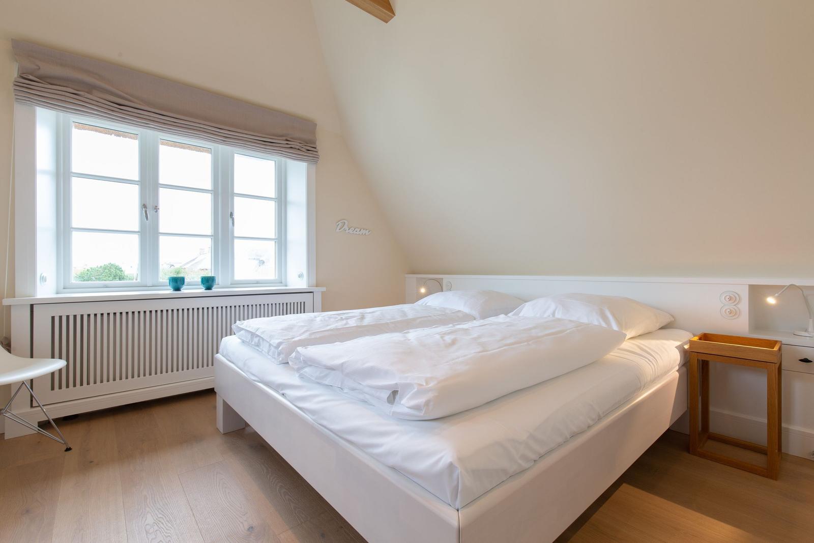 Schlafzimmer mit weißem Bett, Holzboden und Fenster mit Blick nach draußen.