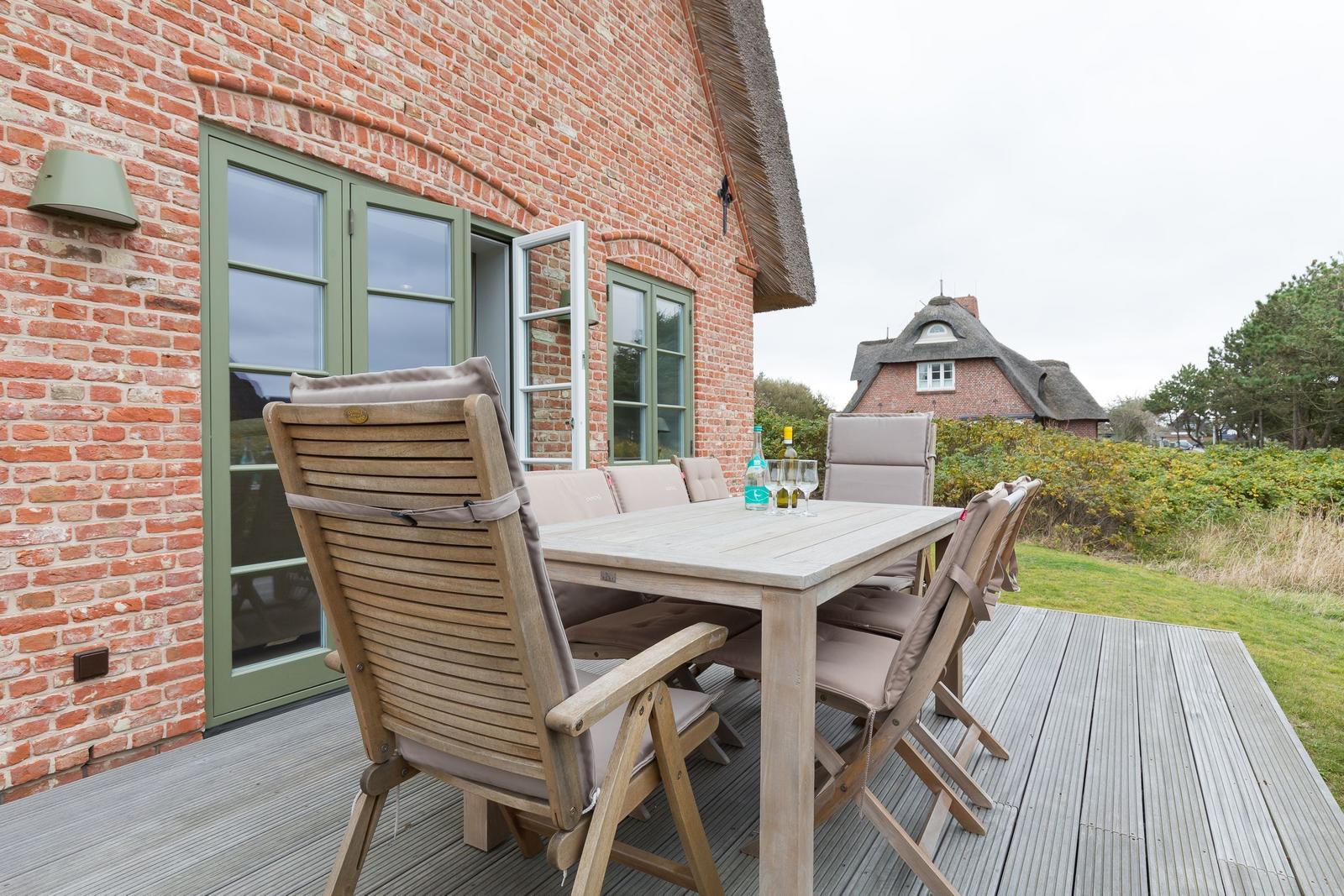 Terrasse mit Holztisch und Stühlen vor einem Backsteinhaus mit grünen Fenstern.
