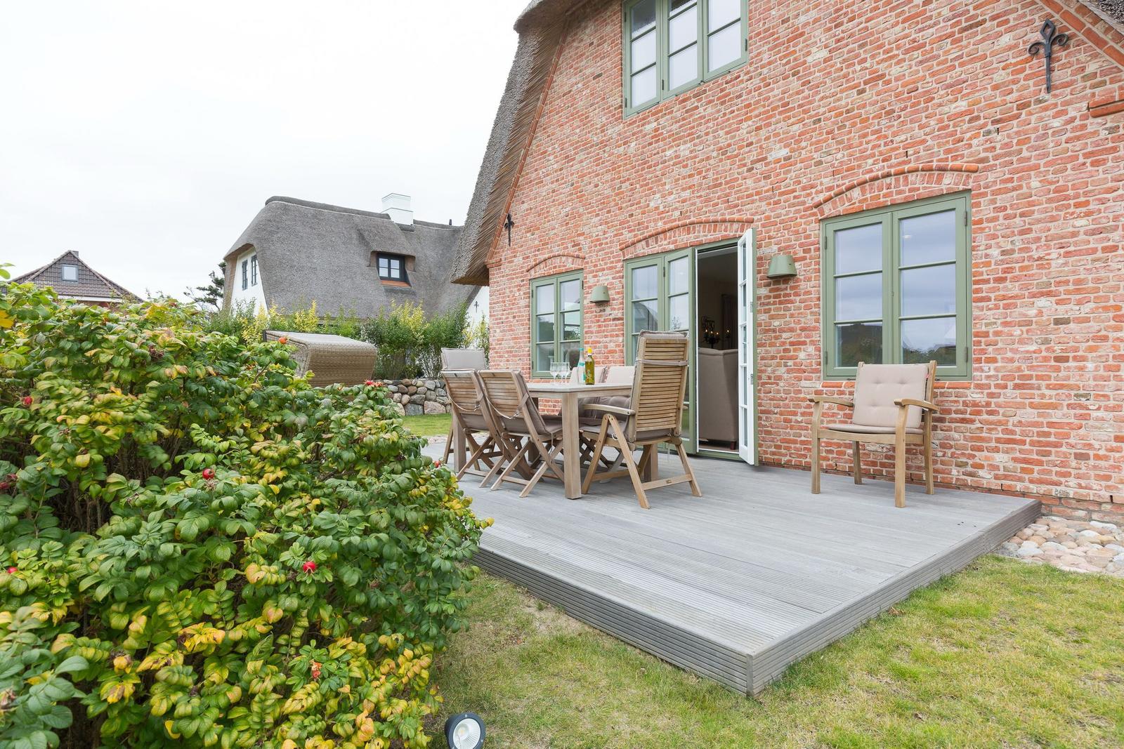 Terrasse mit Tisch und Stühlen an Backsteinhaus mit grünen Fenstern.