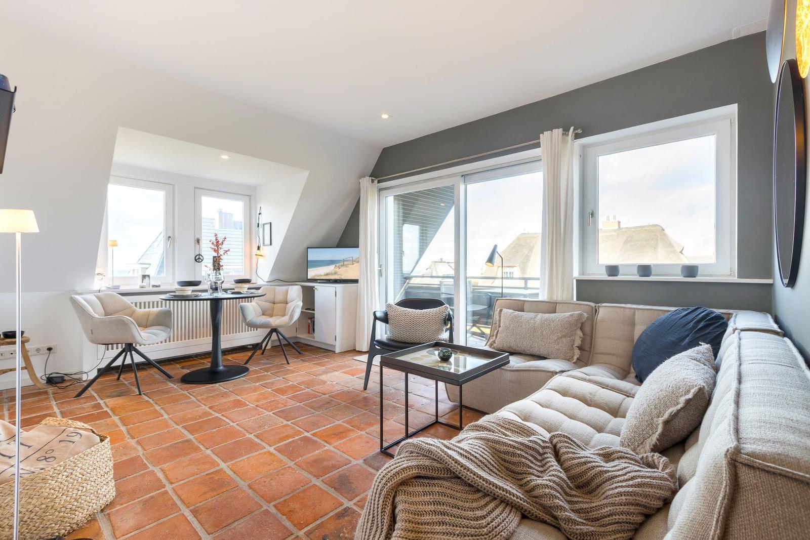 Wohnbereich mit Sofa, Tisch und Fenstern. Terrasse und Blick auf Dächer.