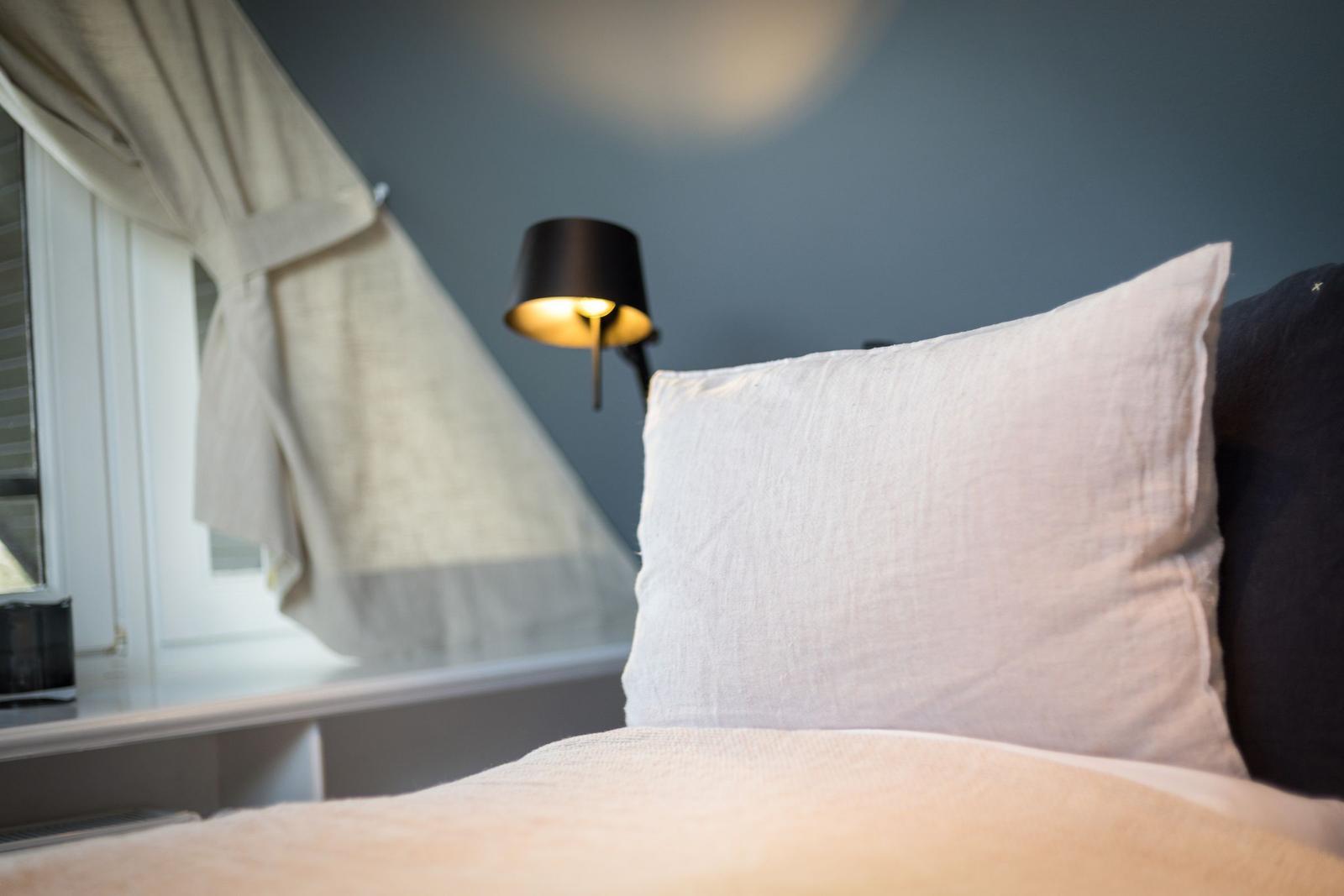 Bettkopf mit Kissen, Wandlampe und Fenster mit Vorhang.
