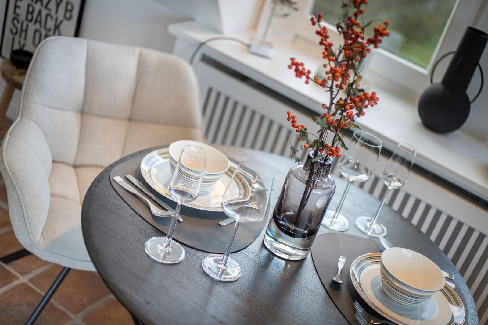Runder Tisch mit Geschirr, Gläsern und Blumenarrangement vor Fenster.