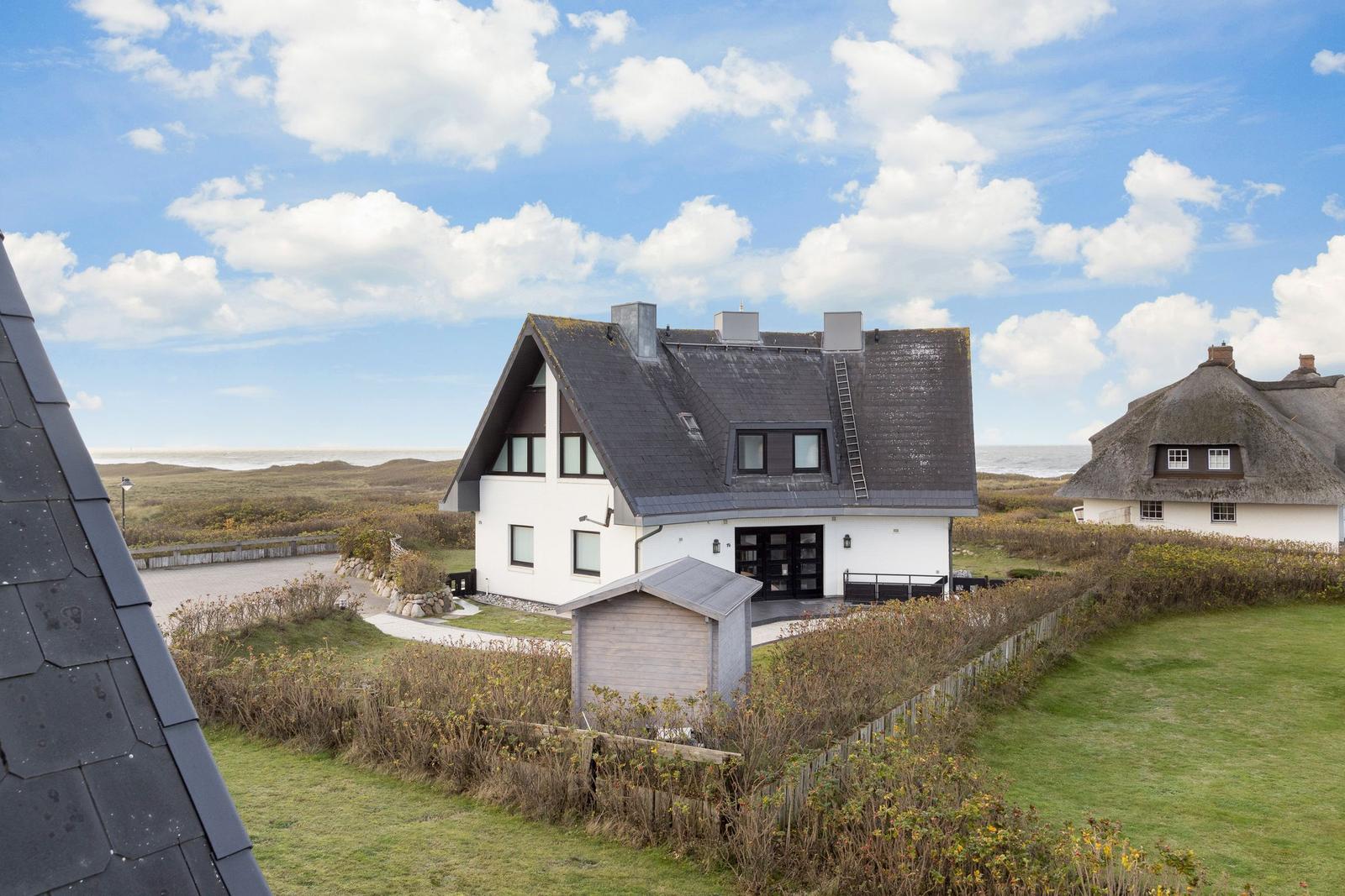 Modernes Haus mit dunklem Dach und weißer Fassade, umgeben von Grünflächen und Strand.
