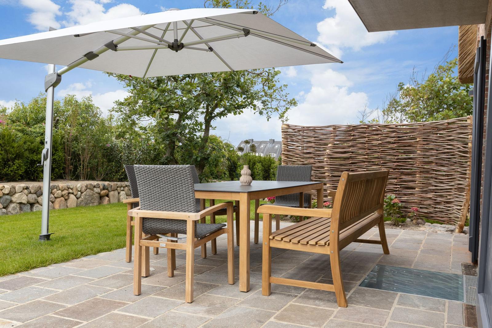 Terrace with table, chairs, and umbrella. Background: greenery and wicker fence.