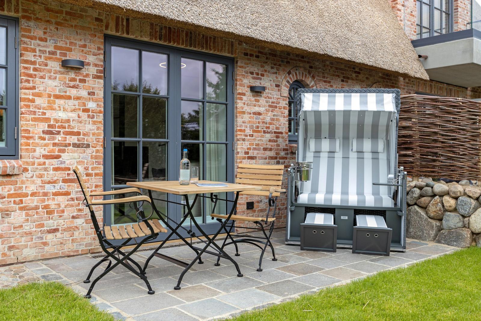 Terrace with table, chairs, and beach chair in front of brick house with thatched roof.
