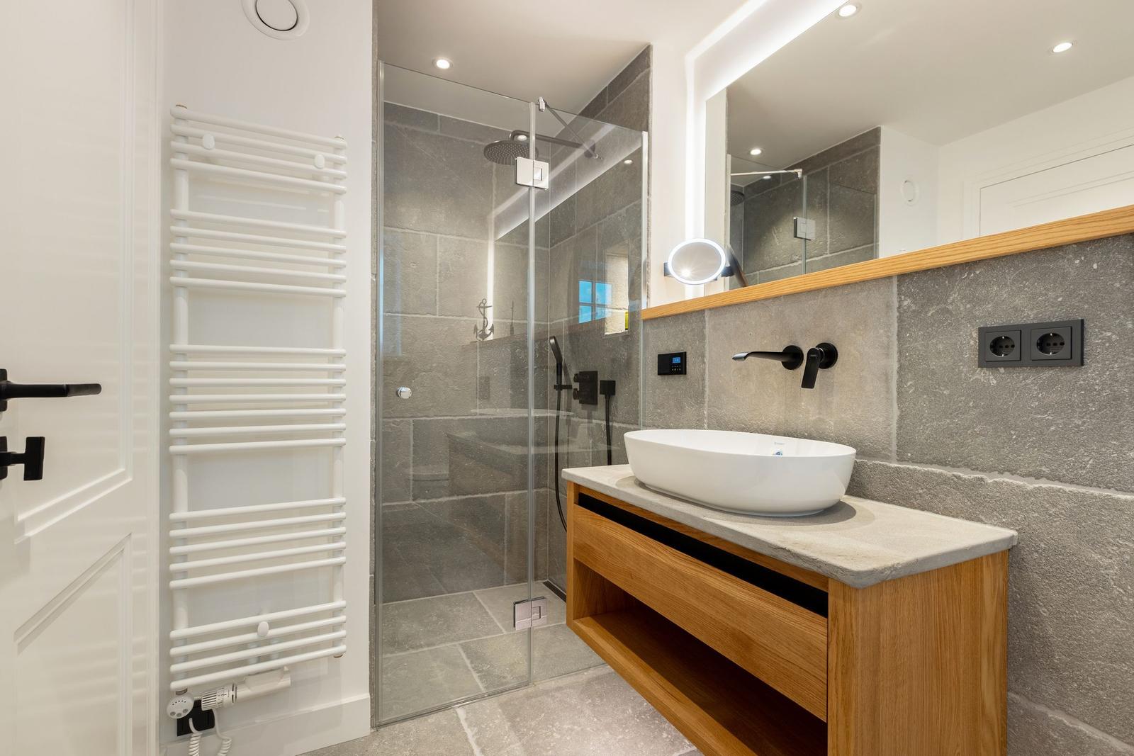Modern shower with glass enclosure, sink with mirror and towel heater on wall.