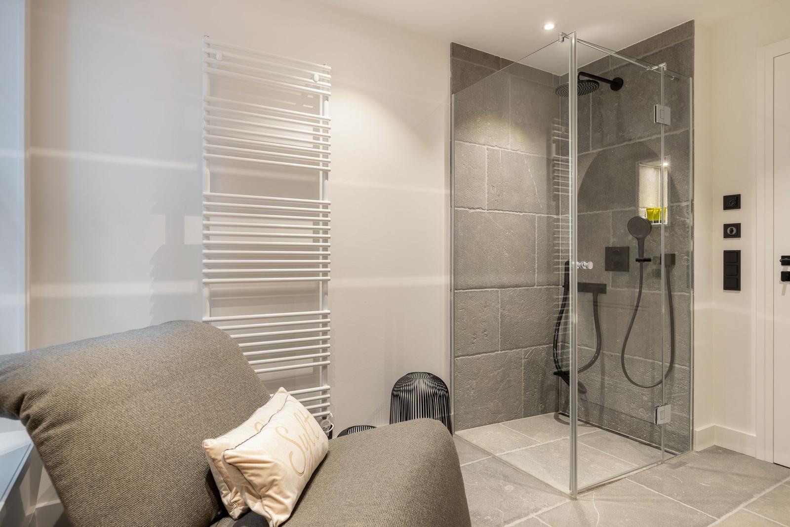 Shower with glass enclosure, showerhead, and handheld shower. Next to a gray chair with pillow.