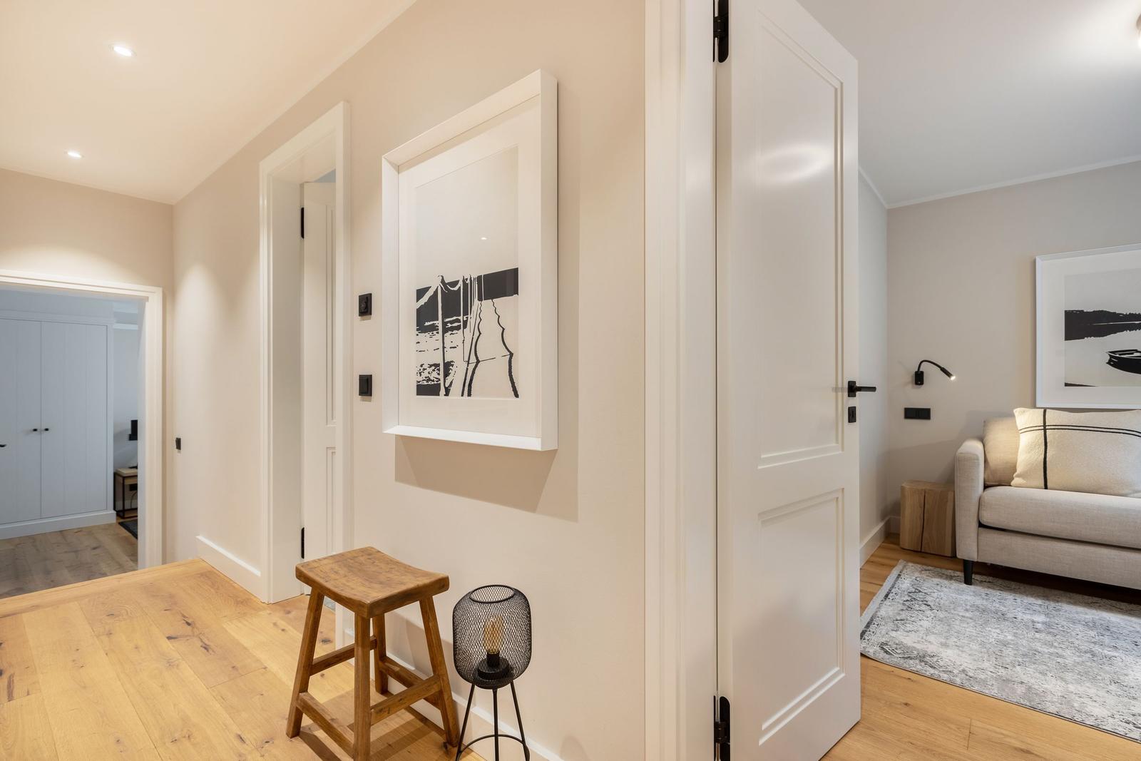 Hallway with wooden floor, stool, and artwork. View into living room with sofa and rug.