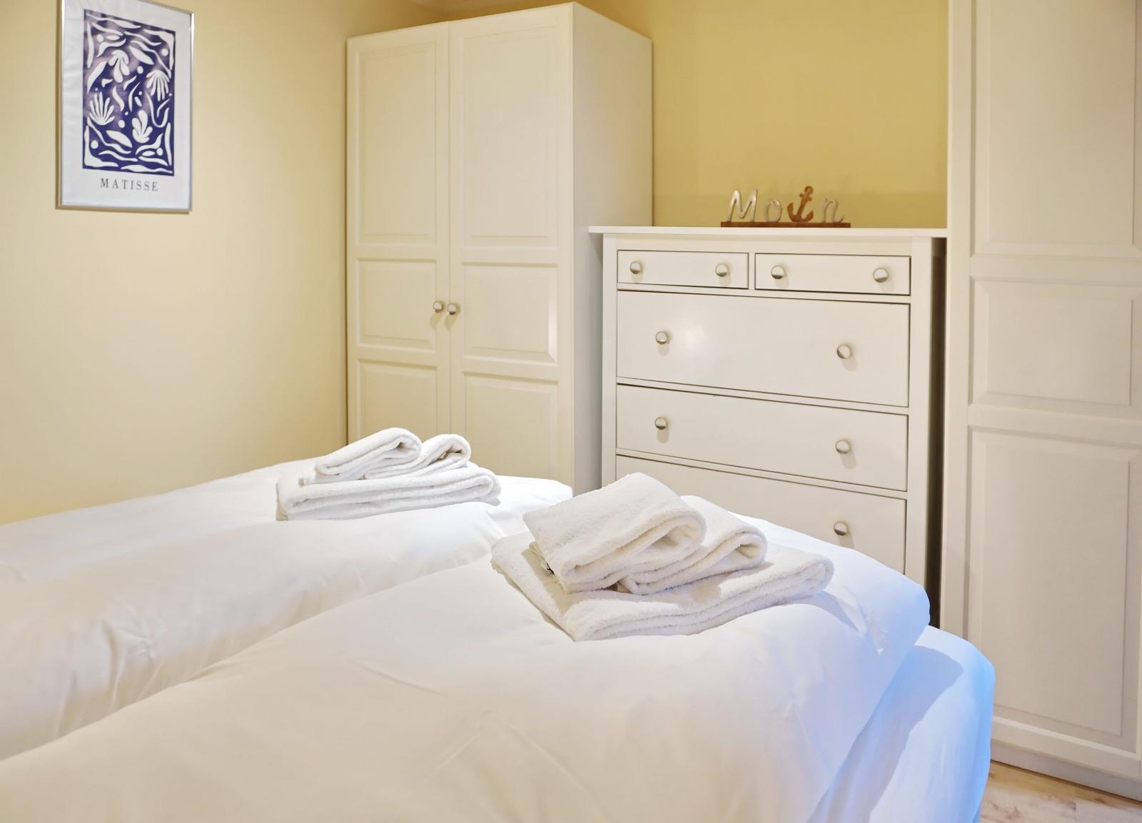 Two beds with white linens and towels. White wardrobe and dresser. Wall art by Matisse.