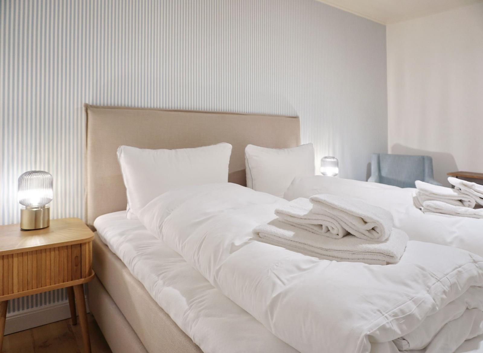 Bed with white linens and folded towels. Wooden nightstand with lamp. Striped wall.