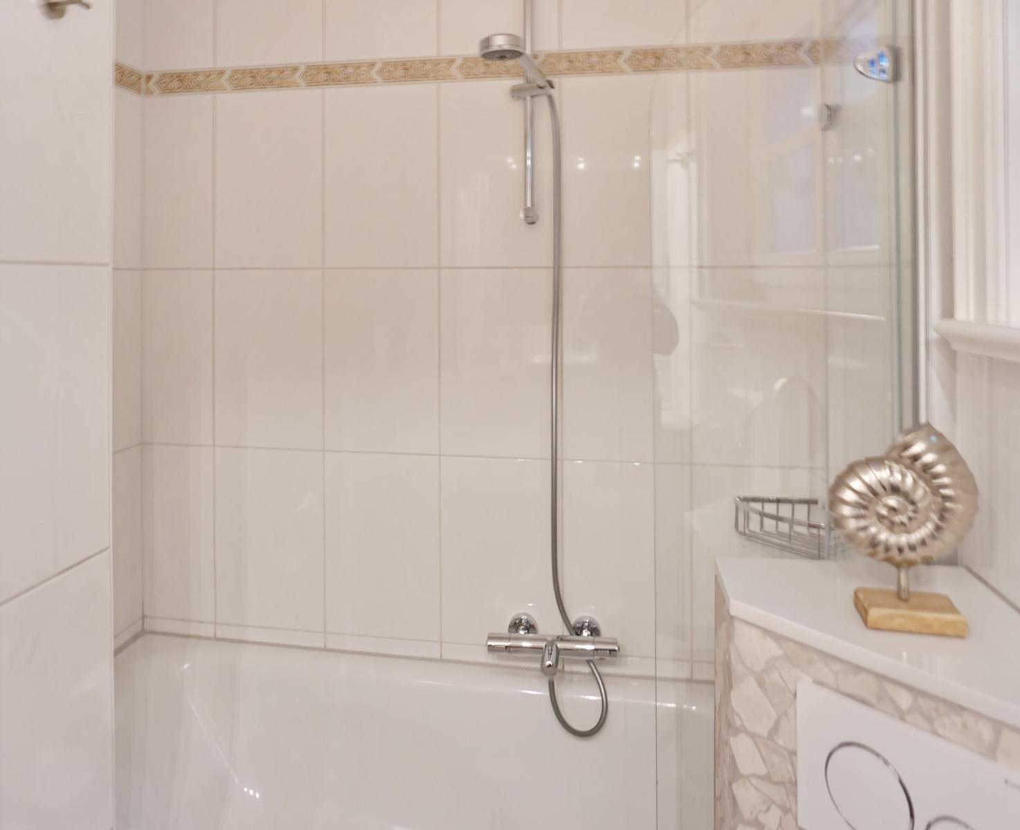 Bath with shower, white tiles, and glass wall. On the sink is a golden seashell figurine.