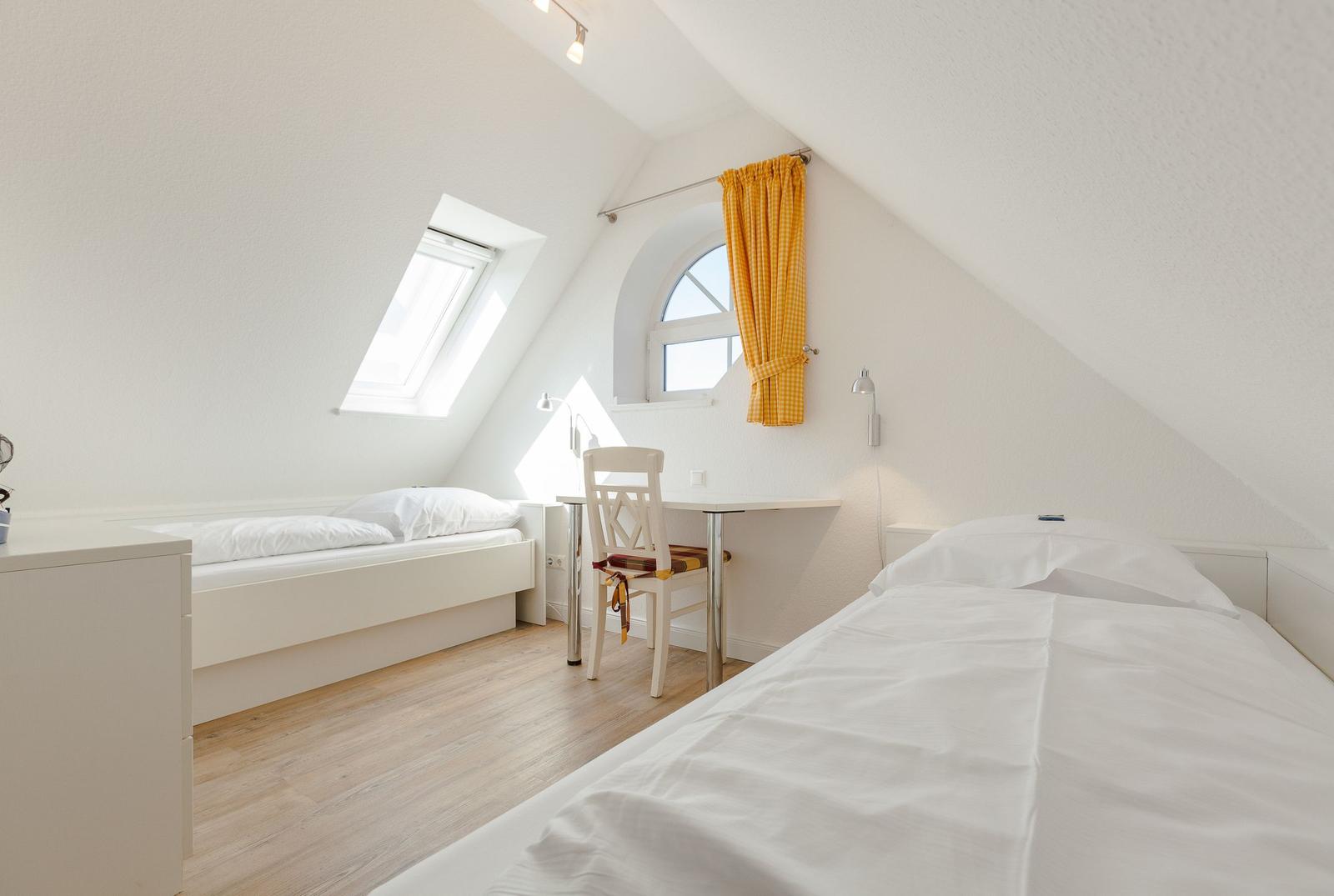 Room with sloped ceiling, two beds, desk, and windows.