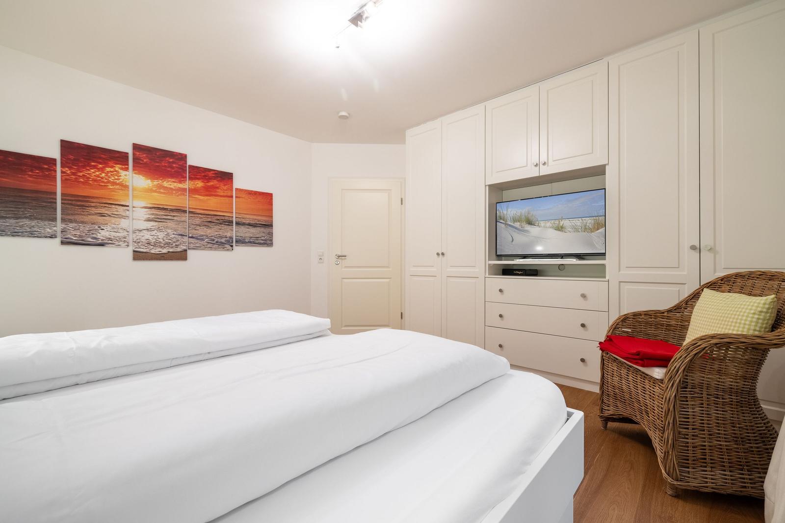 Bedroom with white bed, built-in TV, and wicker chair