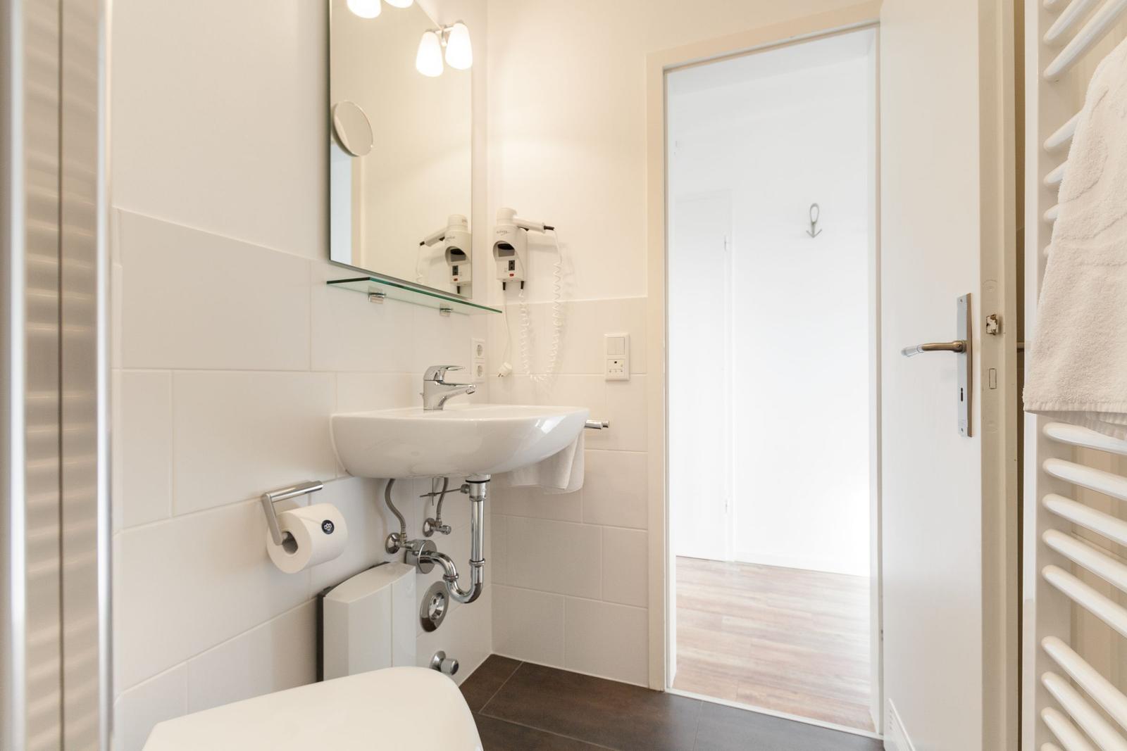 Badezimmer mit Waschbecken, Spiegel und Toilette. Tür führt in Duschbereich.