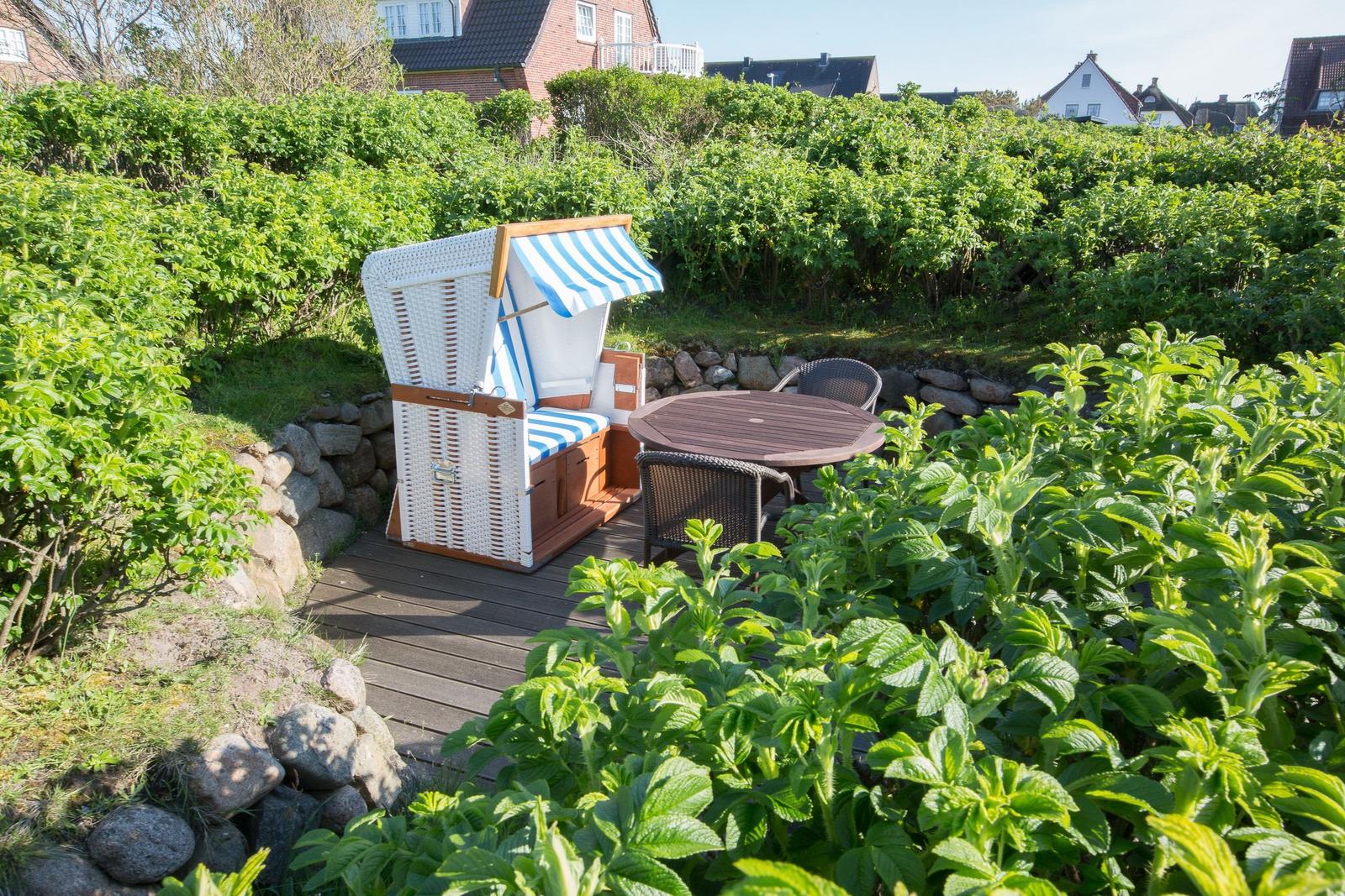 Gartenbereich mit Strandkorb, Tisch und Stühlen. Umgeben von Grünflächen und Steinmauern.