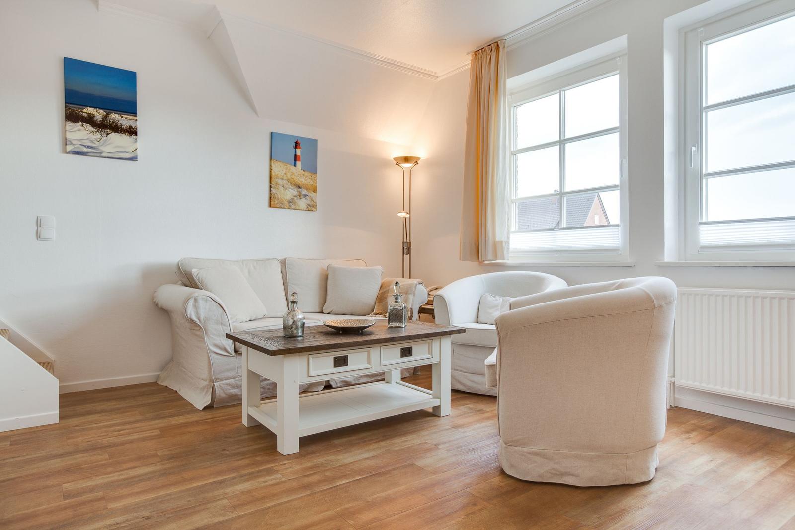 Wohnzimmer mit Sofa, Sessel und Tisch. An den Wänden hängen Bilder. Durch Fenster fällt Licht.
