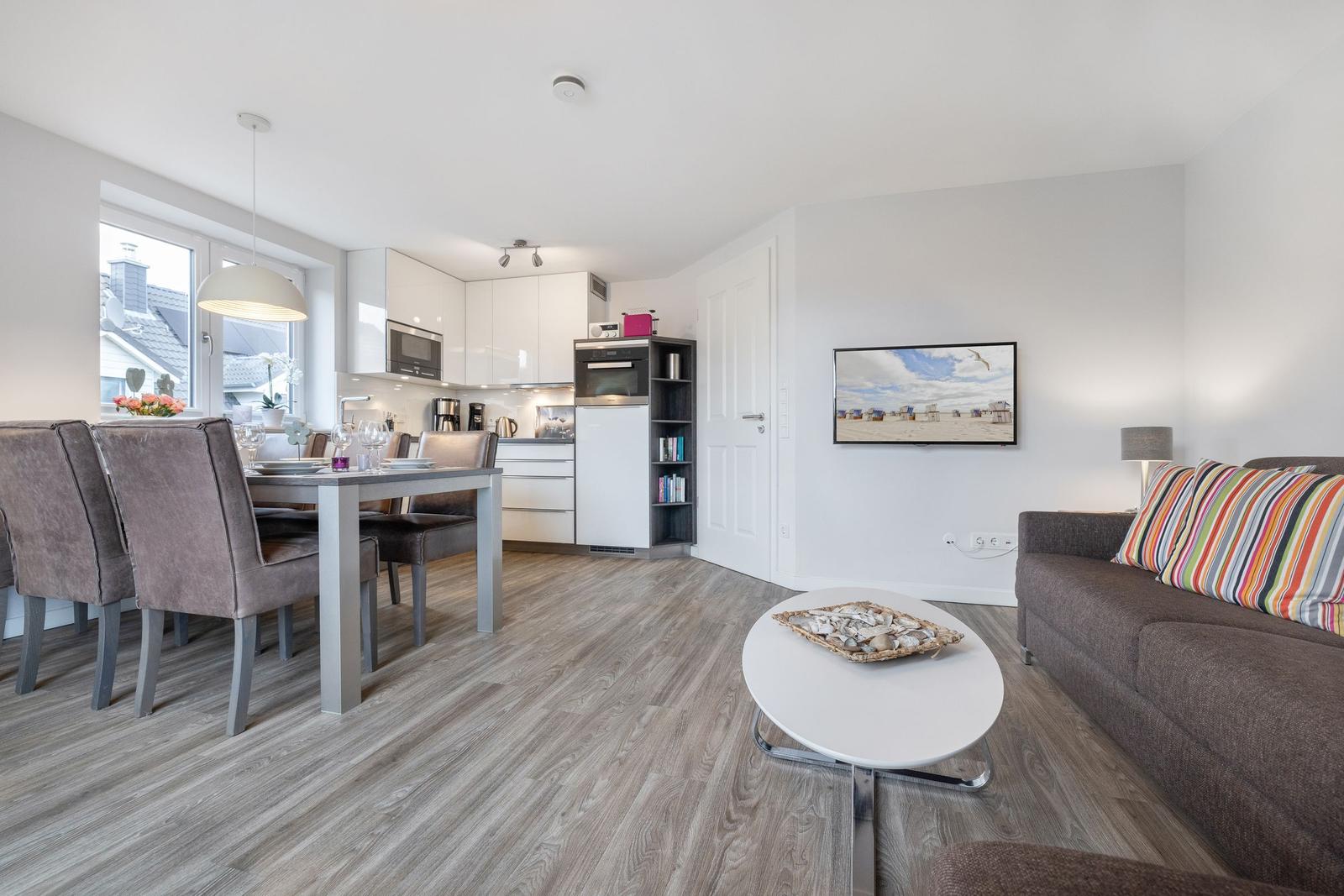 Living kitchen with dining table, sofa, and TV. Wooden floor and white kitchen area.
