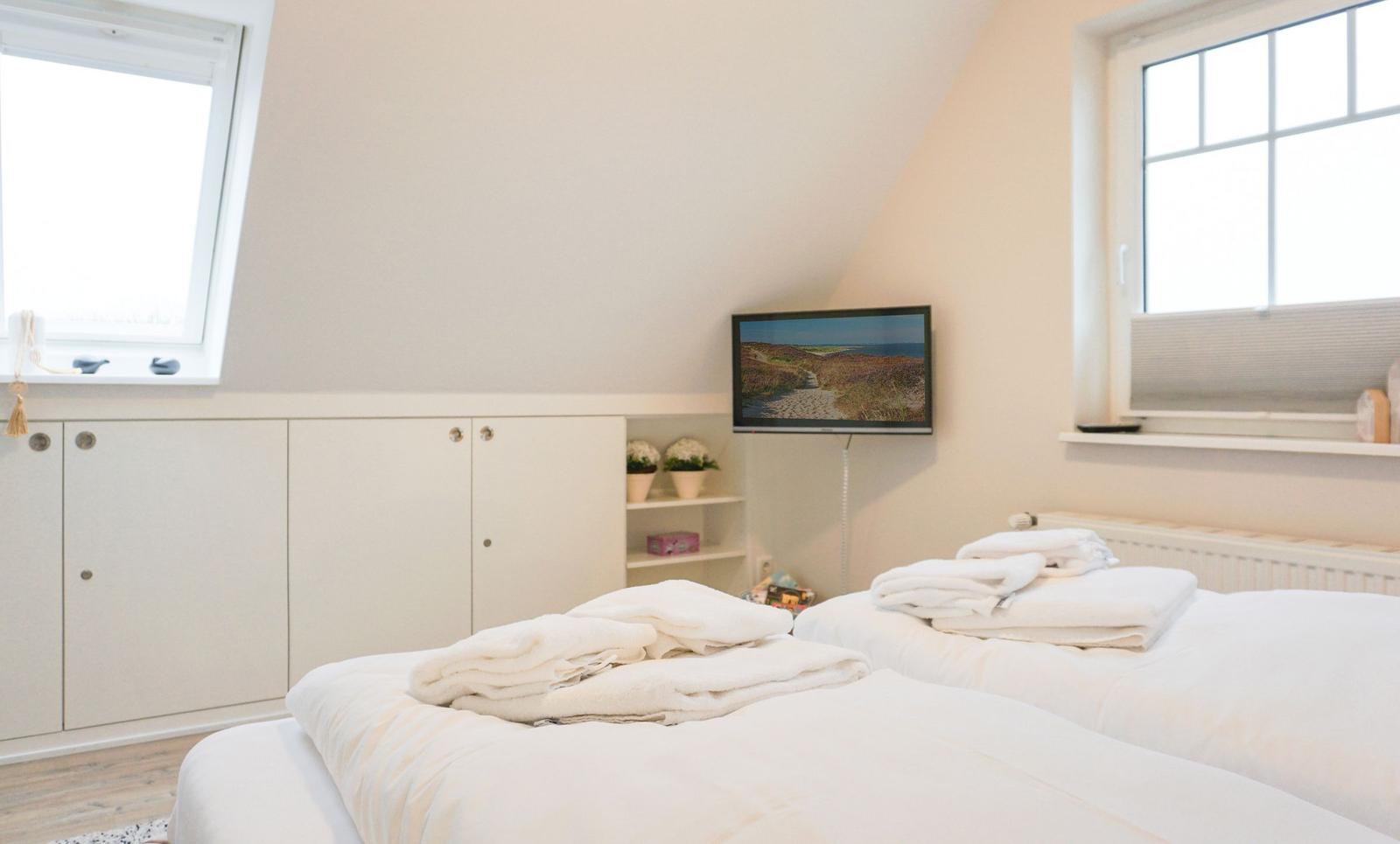 Two beds with white sheets, TV, and windows in a bedroom.