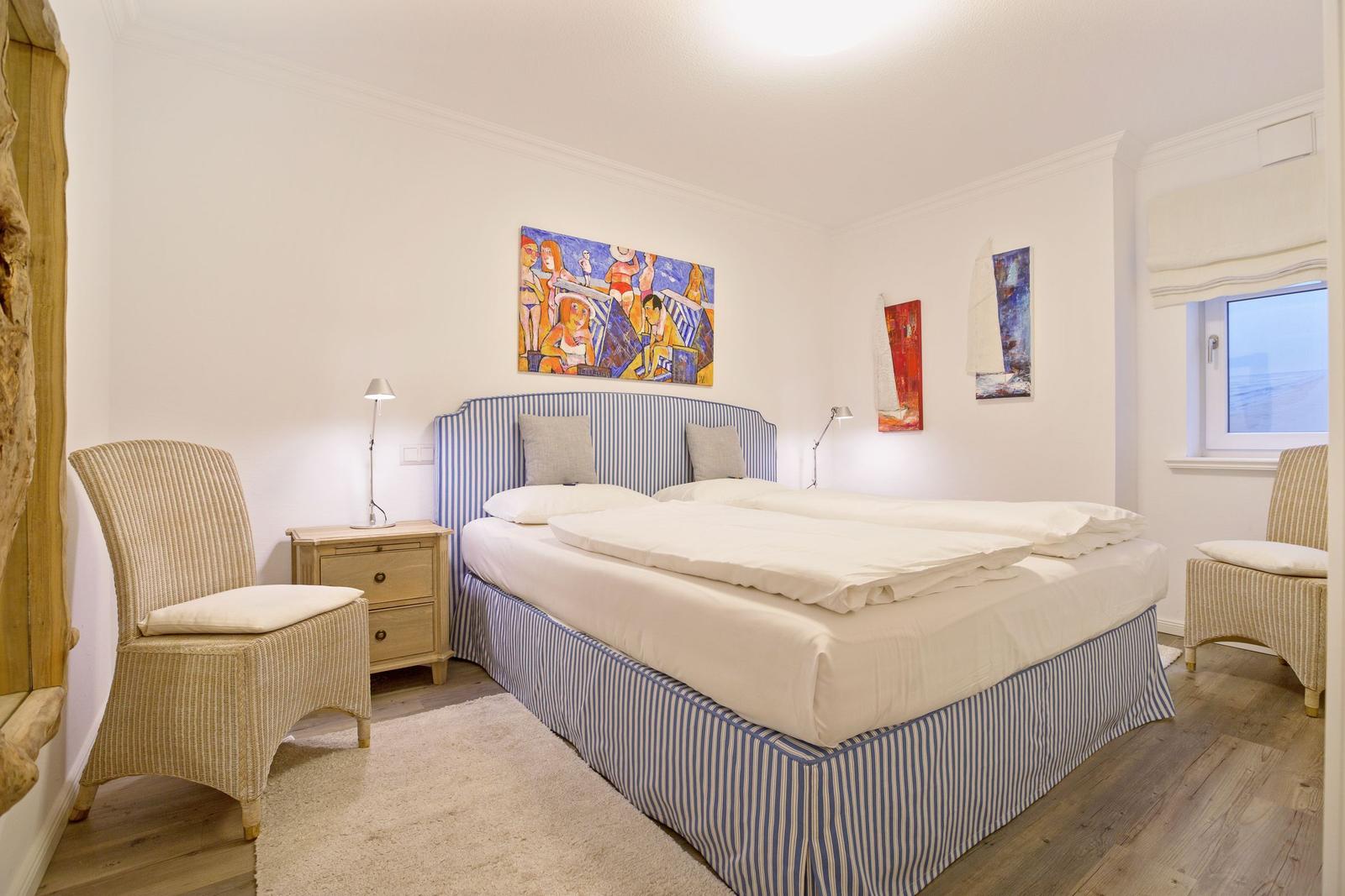 Schlafzimmer mit Bett, Nachttisch und Stühlen. Wandbild und Fenster sichtbar.