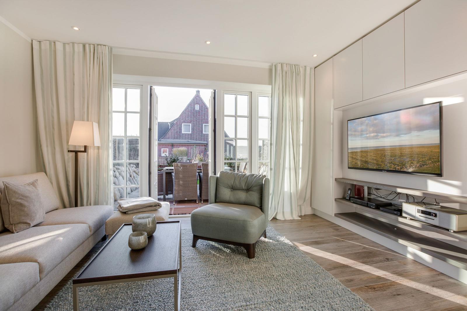 Wohnzimmer mit Sofa, Sessel, Couchtisch und TV. Durchgang zur Terrasse mit Blick auf Haus.