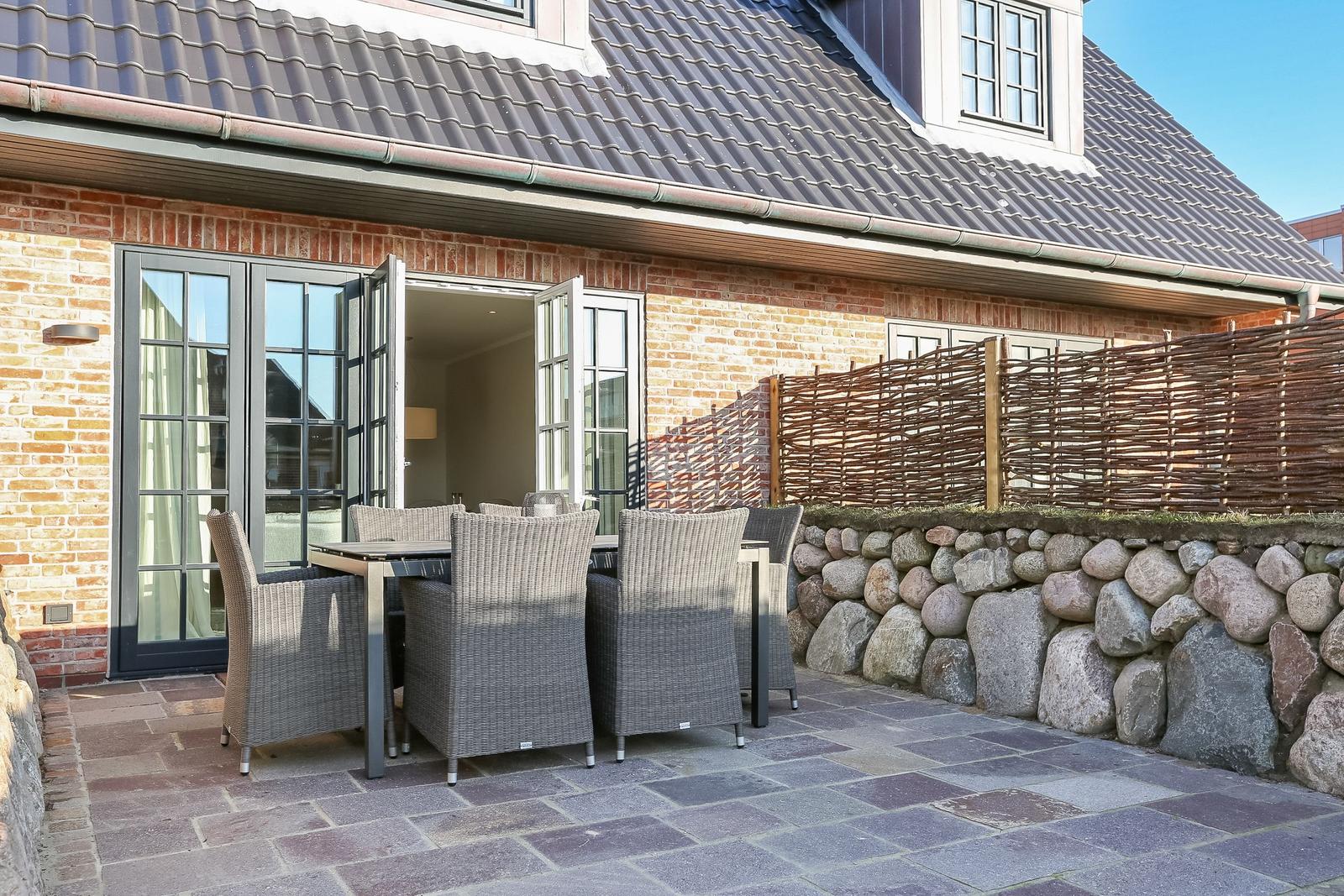 Terrasse mit Tisch und Stühlen vor einem Haus mit Steinmauer und Holzzaun.