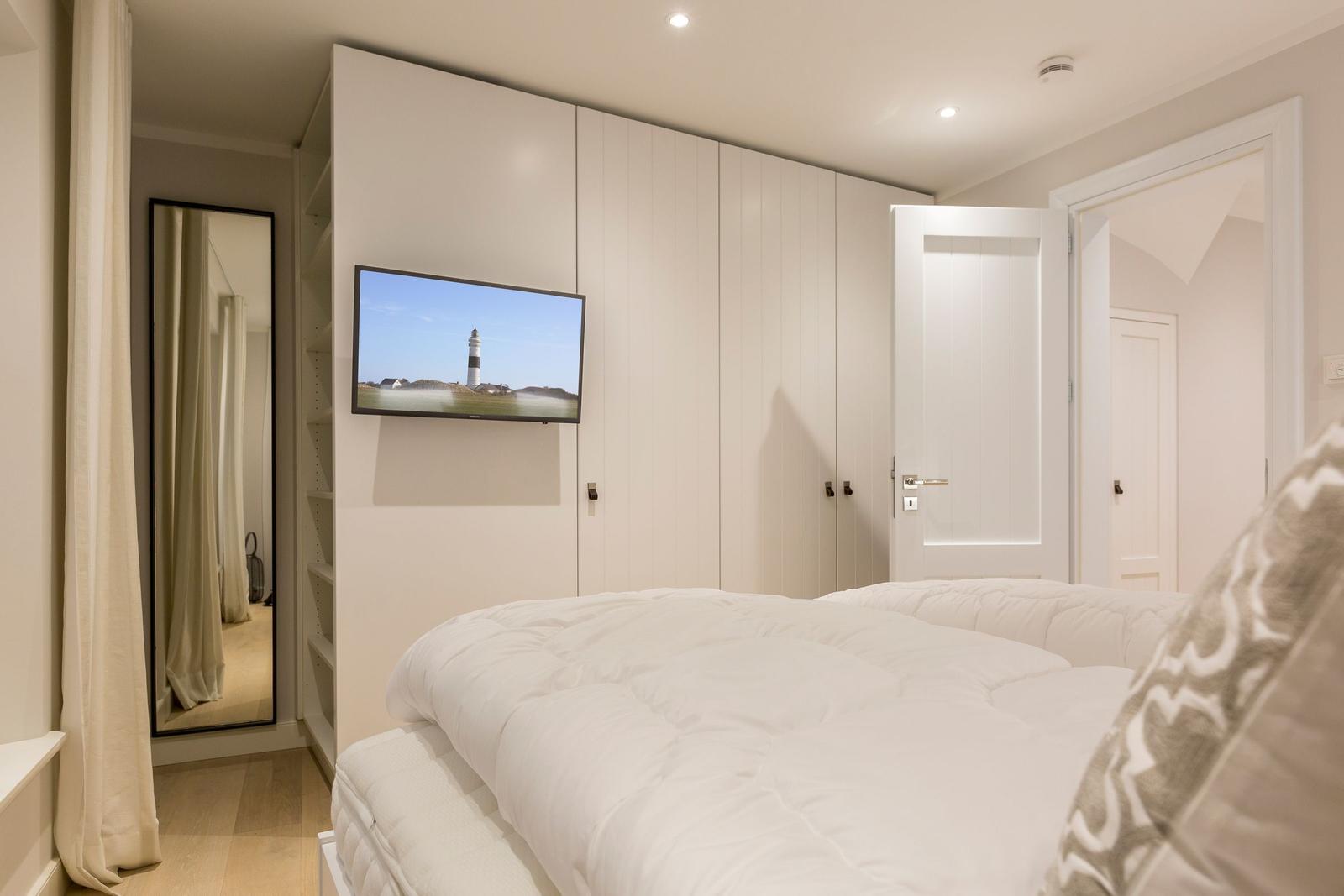 Schlafzimmer mit weißem Bett, Kleiderschrank und Fernseher an der Wand.