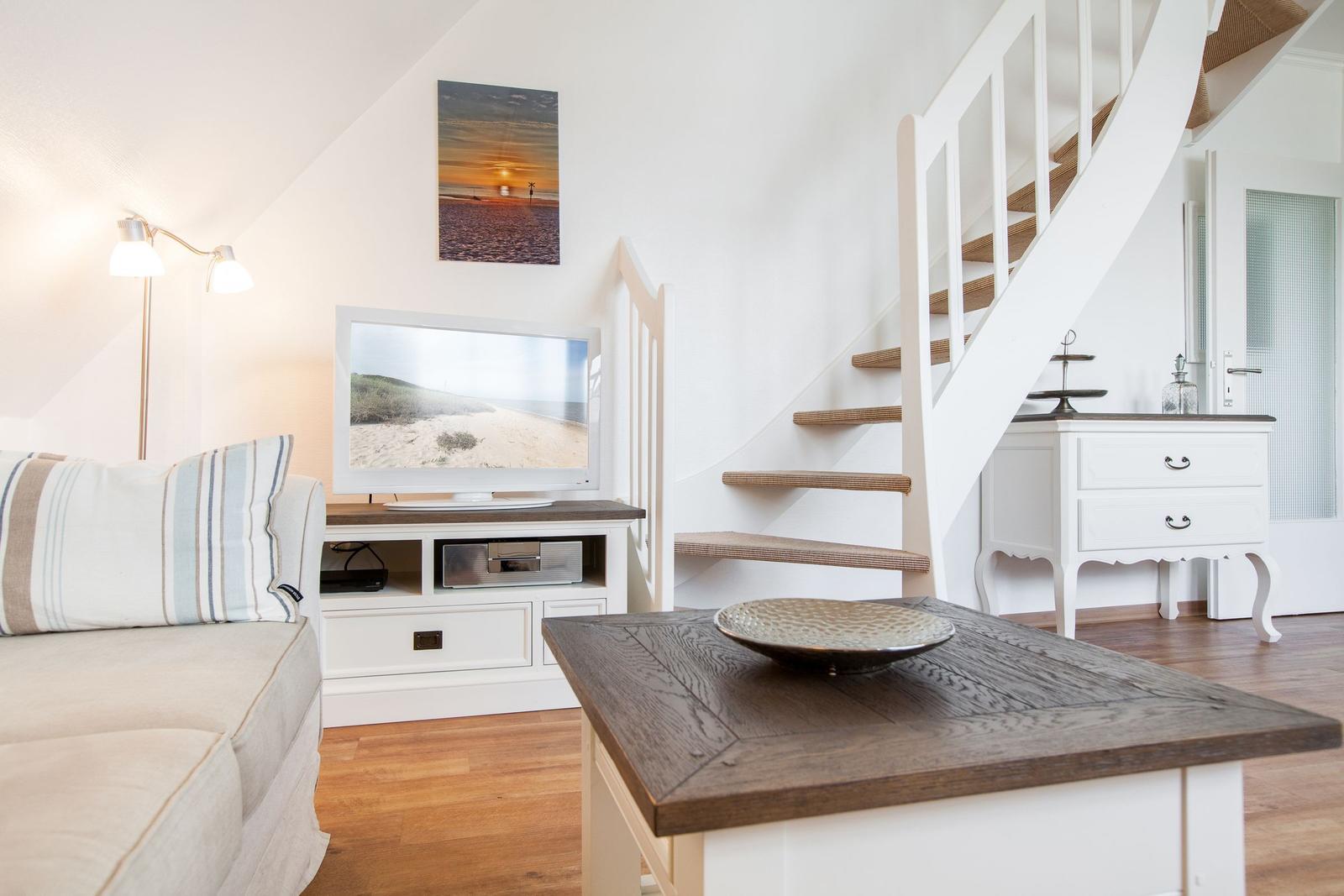 Living room with TV, sofa, and storage unit. Wooden staircase leads to upper level.