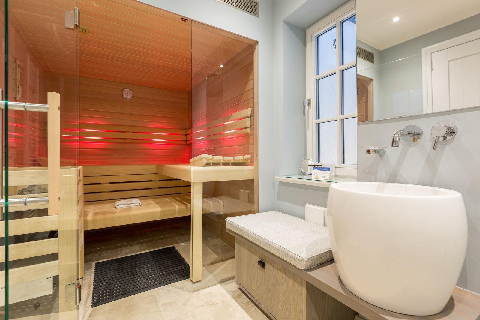 Sauna with wooden bench and red light, beside freestanding tub and window.