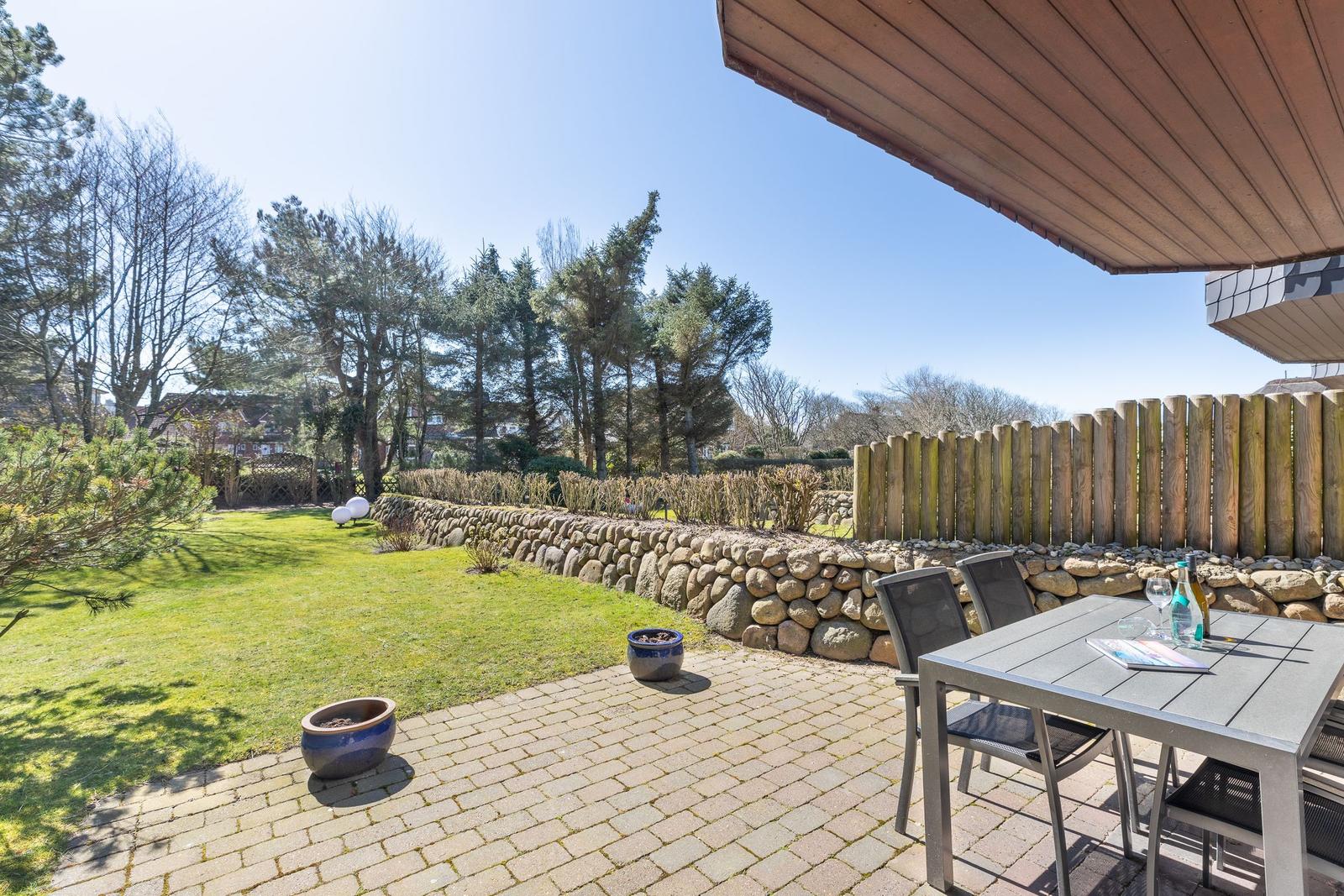 Terrace with table and chairs, paved ground, lawn, and stone wall in background.