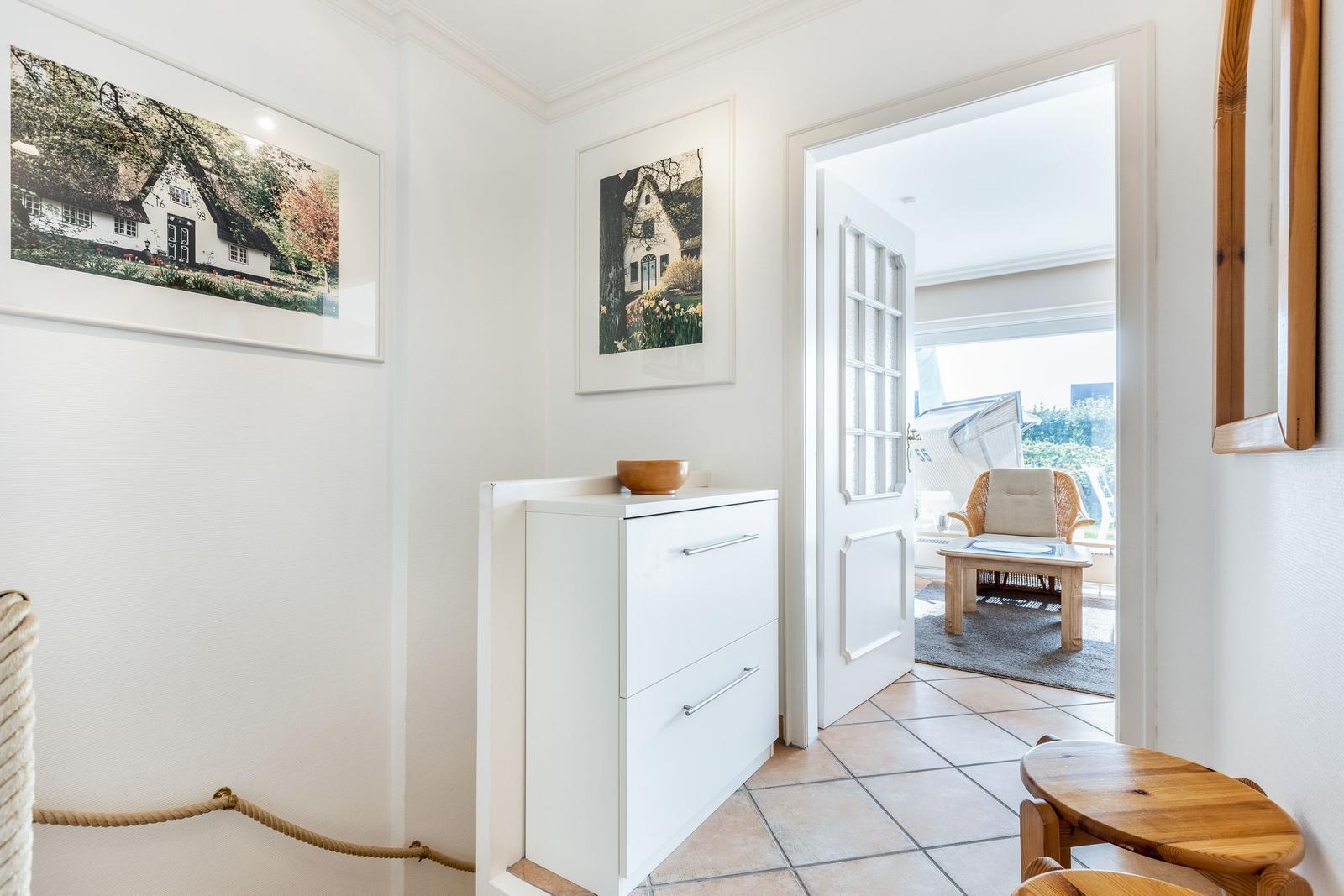 Entrance with white walls, pictures, and a white cabinet. Through door, living area with chair is visible.