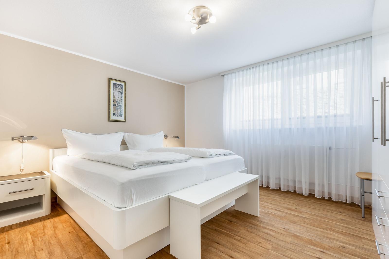 Bedroom with white bed, wooden floor, and large windows with curtains.