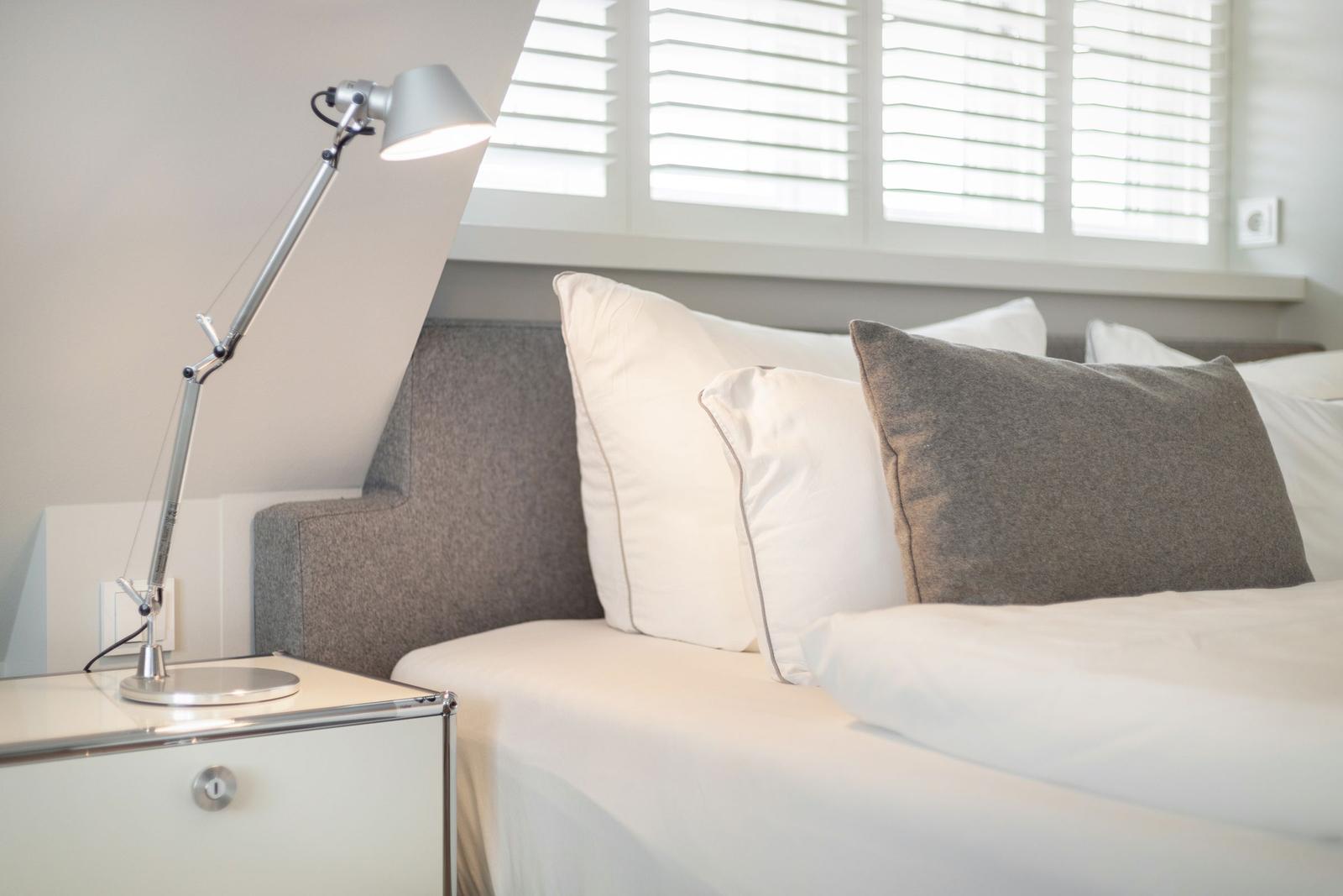Schlafzimmer mit Bett, Nachttisch und Stehlampe unter Fenster mit Jalousie.