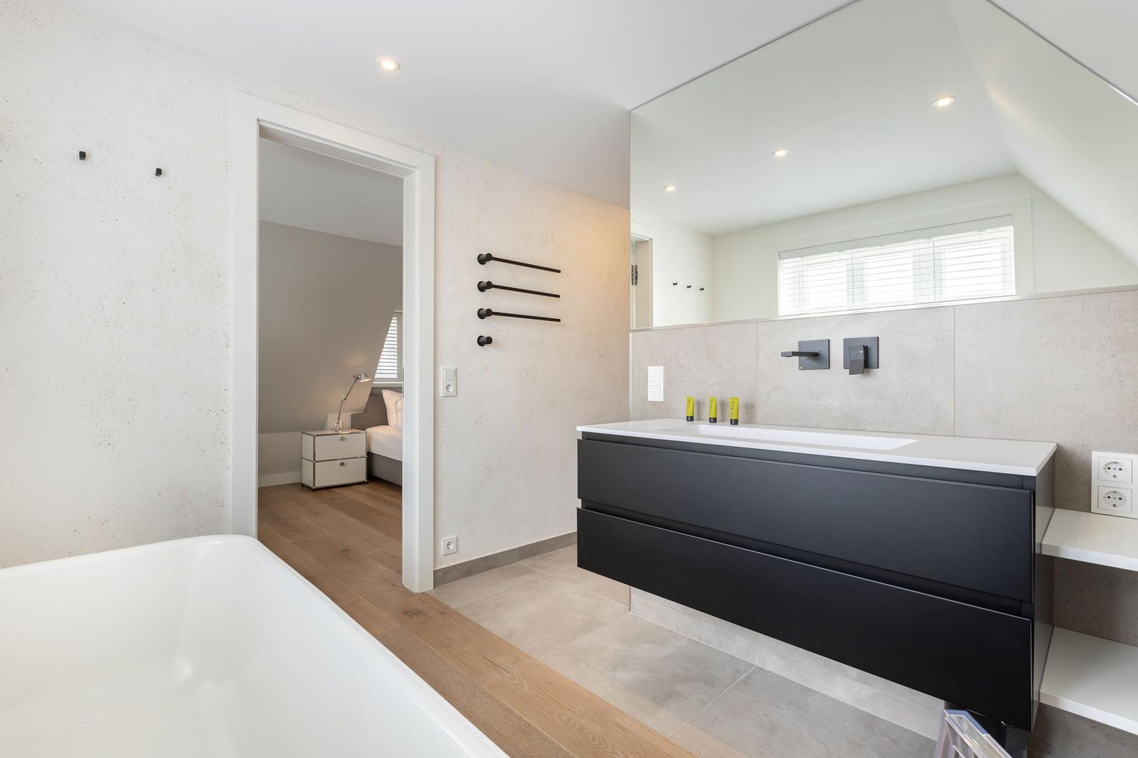 Badezimmer mit Dusche, Waschbecken und Badewanne. Blick in Schlafzimmer mit Bett.