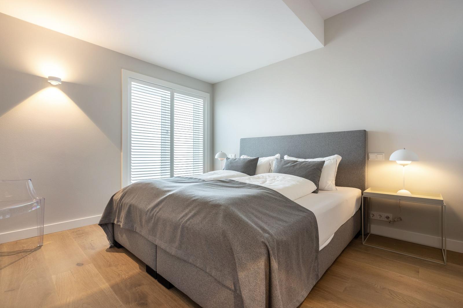 Schlafzimmer mit Bett, Nachttisch und Fenster mit Jalousie.