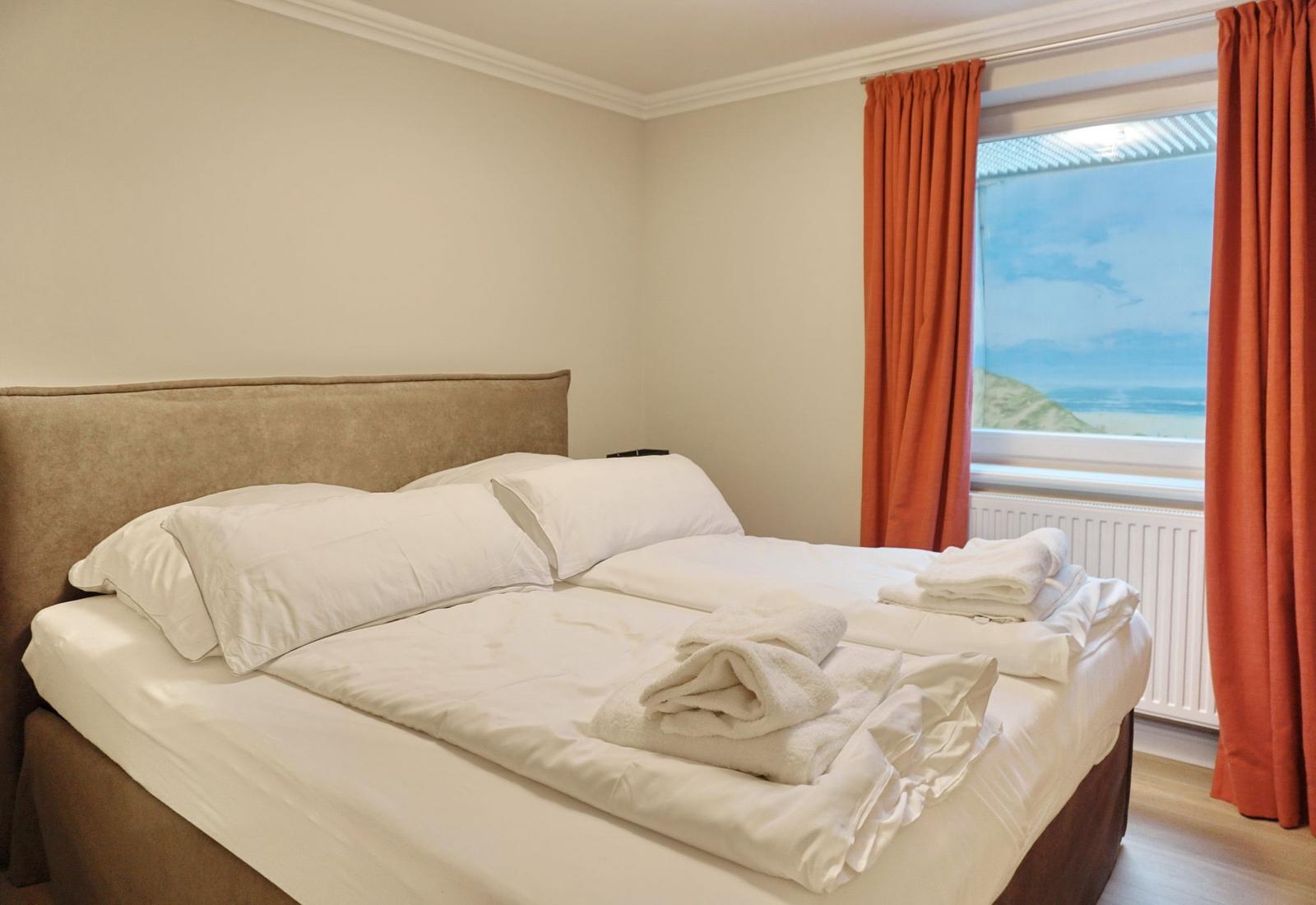 Doppelbett mit weißer Bettwäsche und orangefarbenen Vorhängen vor Fenster mit Meerblick.