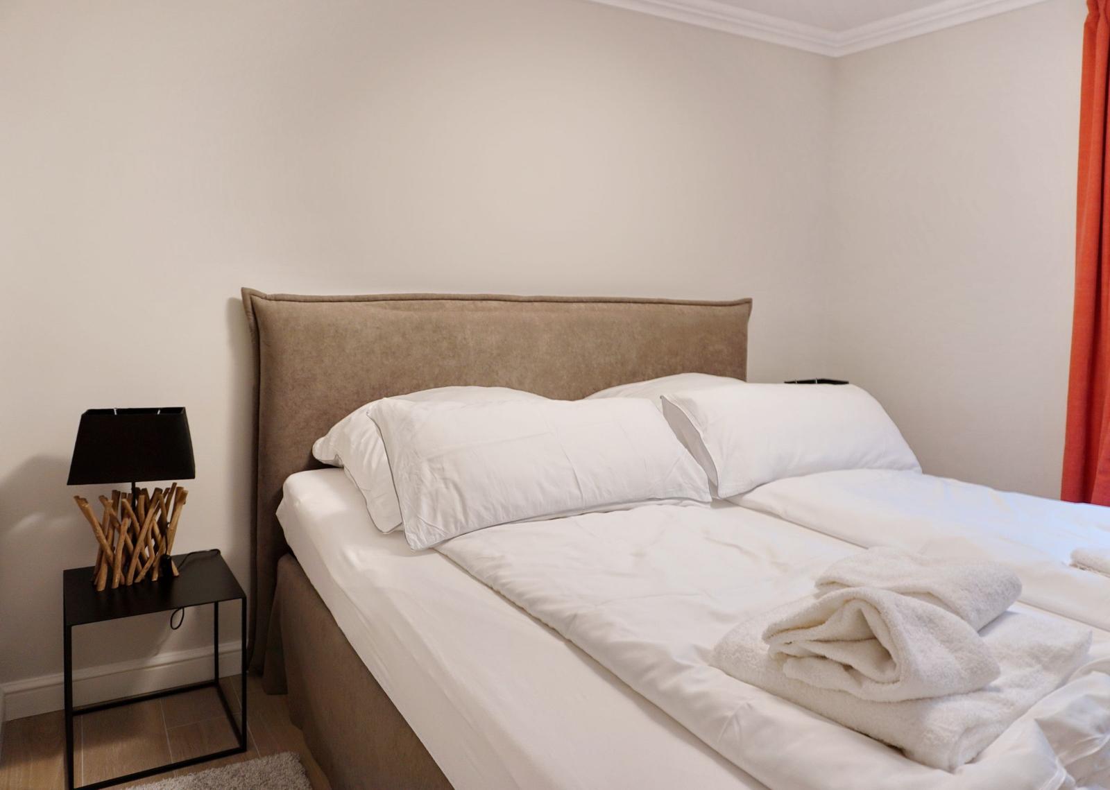 Ein Bett mit weißen Laken und Kissen. Neben dem Bett steht ein Nachttisch mit einer Lampe.