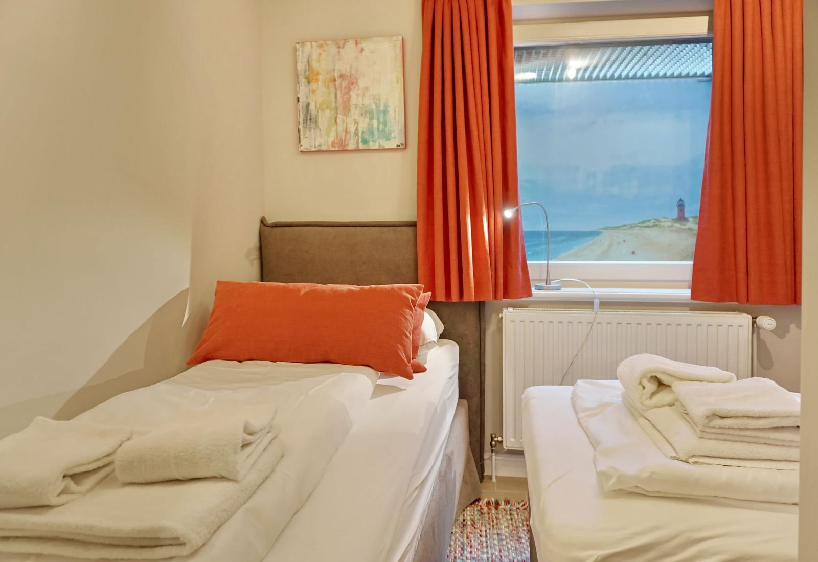 Zwei Betten mit weißen Bettwäsche und orangefarbenen Kissen. Fenster mit orangefarbenen Vorhängen.