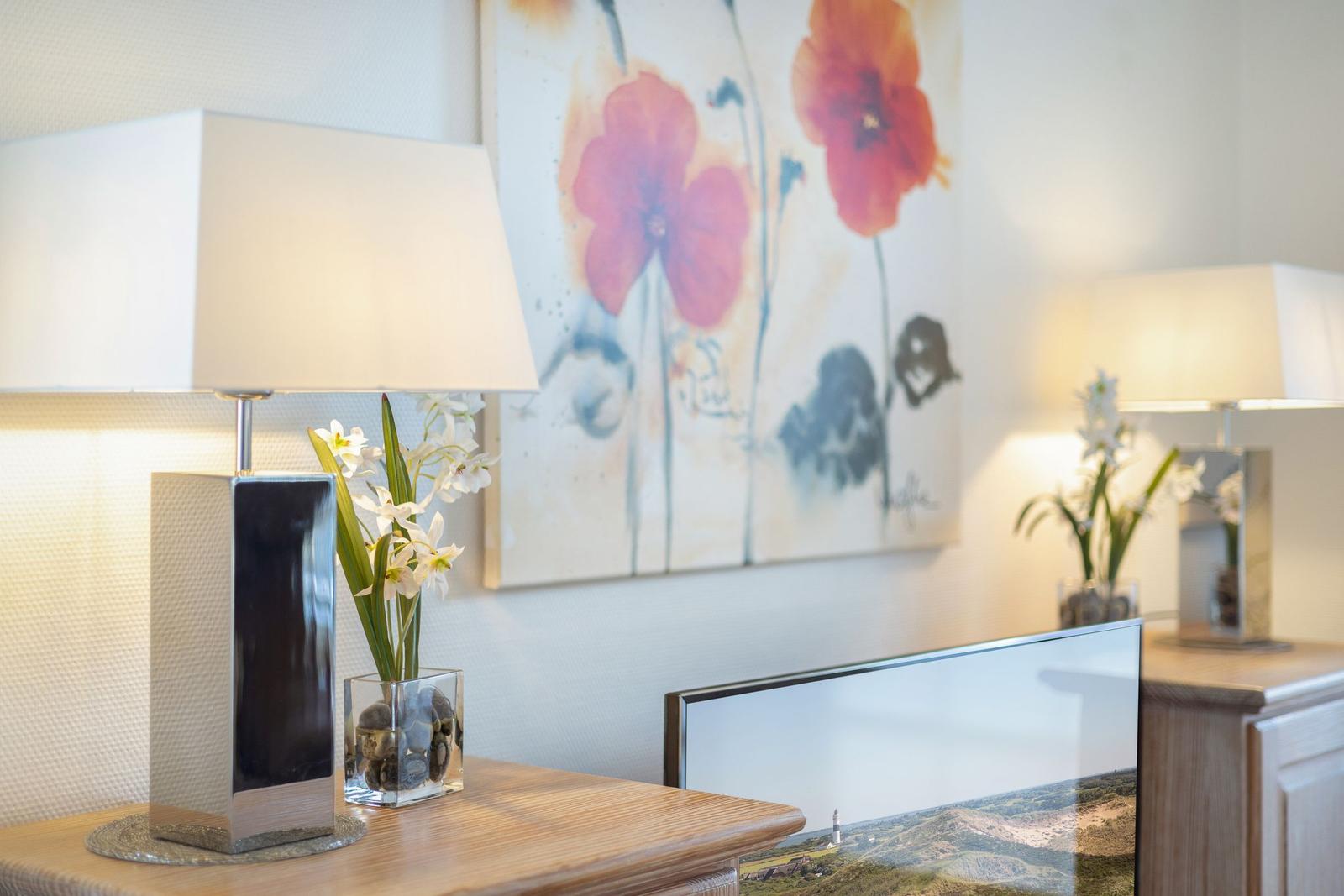 TV on wooden console with lamps and floral arrangement on wall.