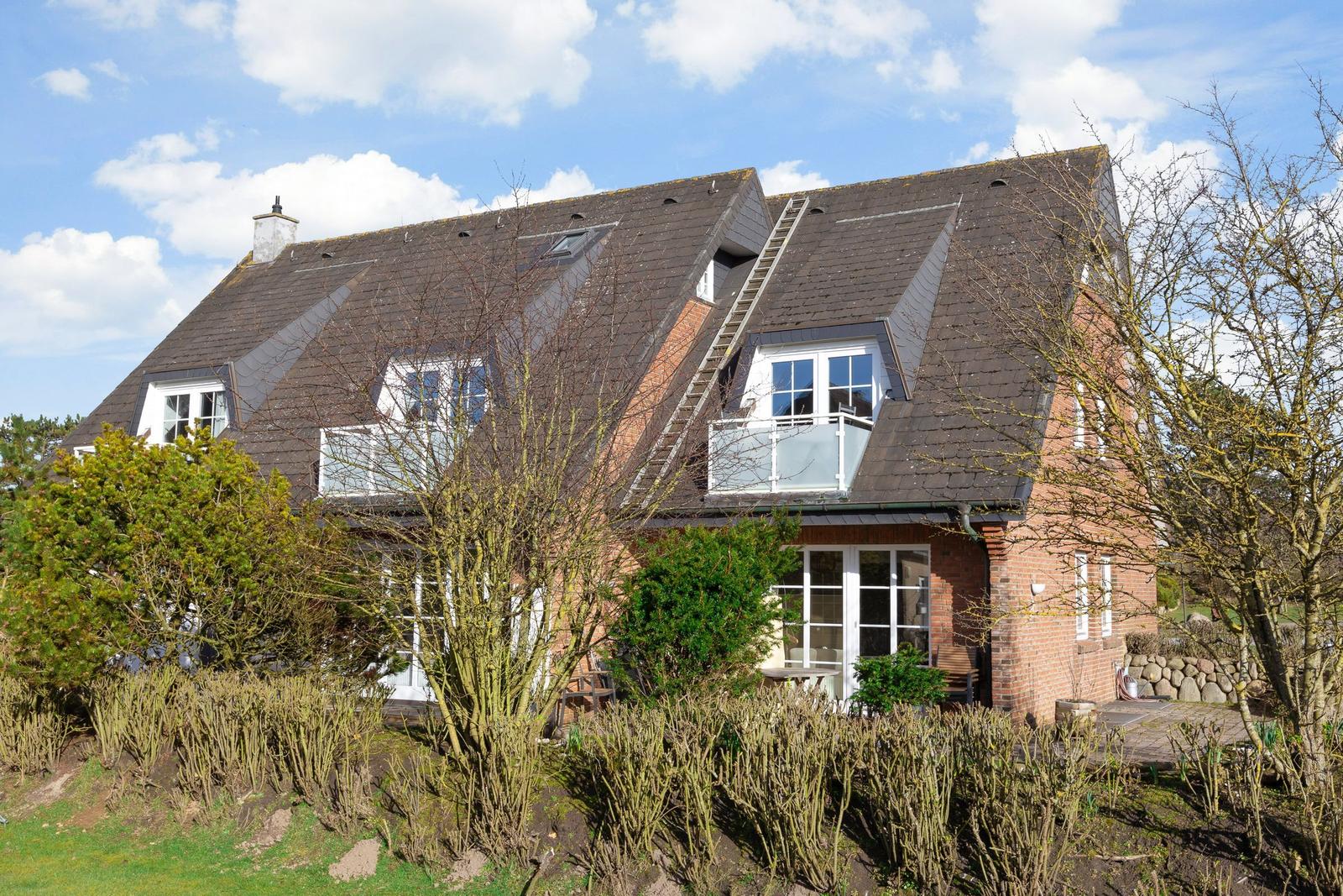 A two-story house with a rooftop terrace and balcony. Surrounded by trees and shrubs.