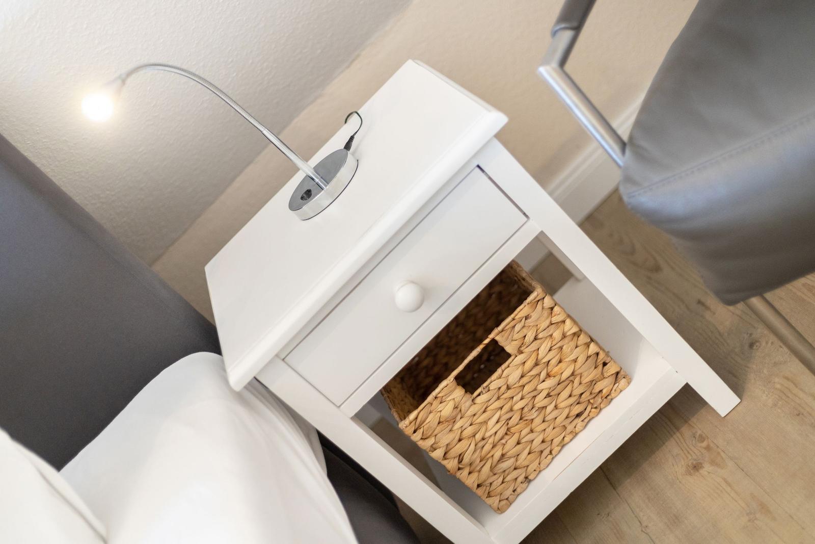 White nightstand with drawer and basket. Lamp and bed are visible.
