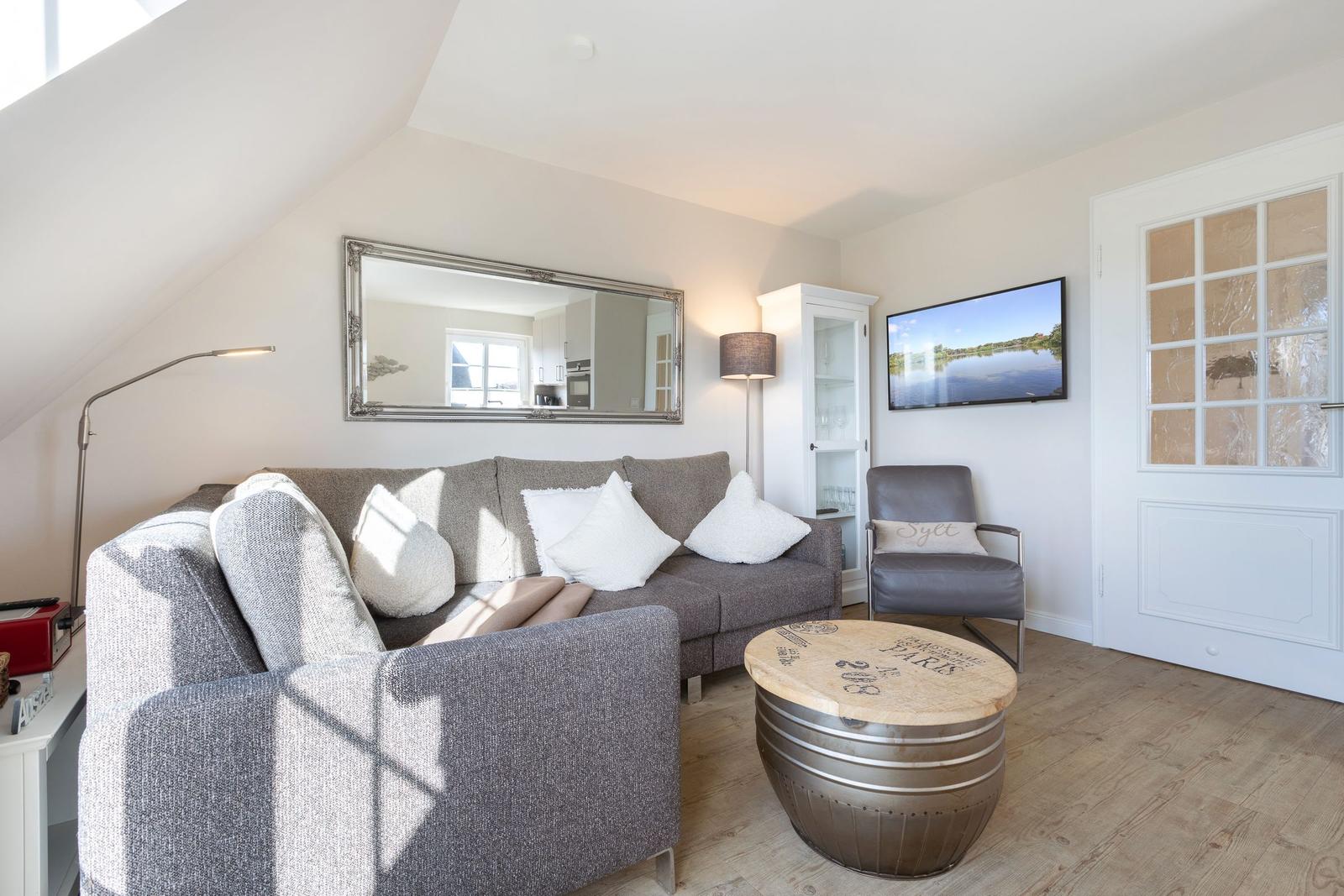 Living room with sofa, armchair, and round table. Mirror and TV on the wall.