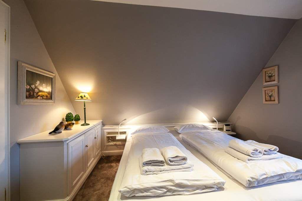 Two beds with white linens and towels in an attic room with lamps and pictures.