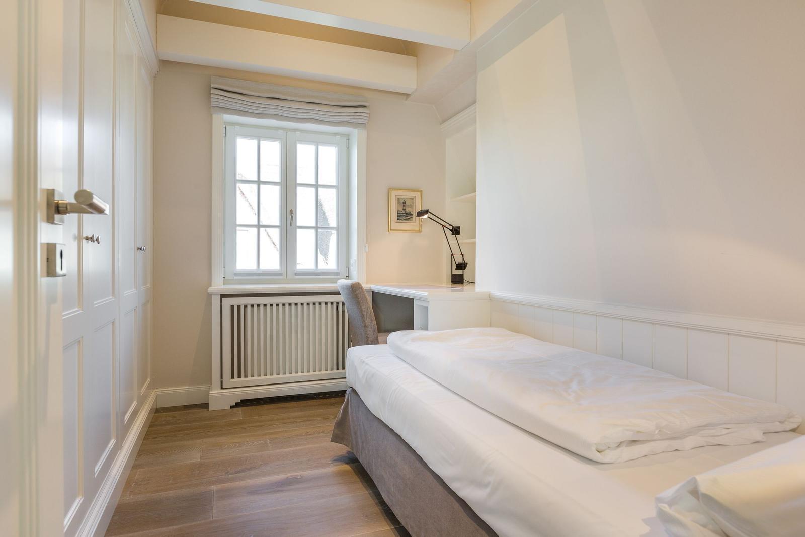 Schlafzimmer mit Bett, Schreibtisch und Fenster. Holzboden und weiße Wände.