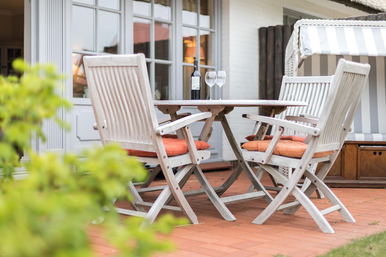 Terrasse mit Tisch, zwei Stühlen und Weinflasche. Hintergrund: Haus mit Fenstern und Sonnenschutz.