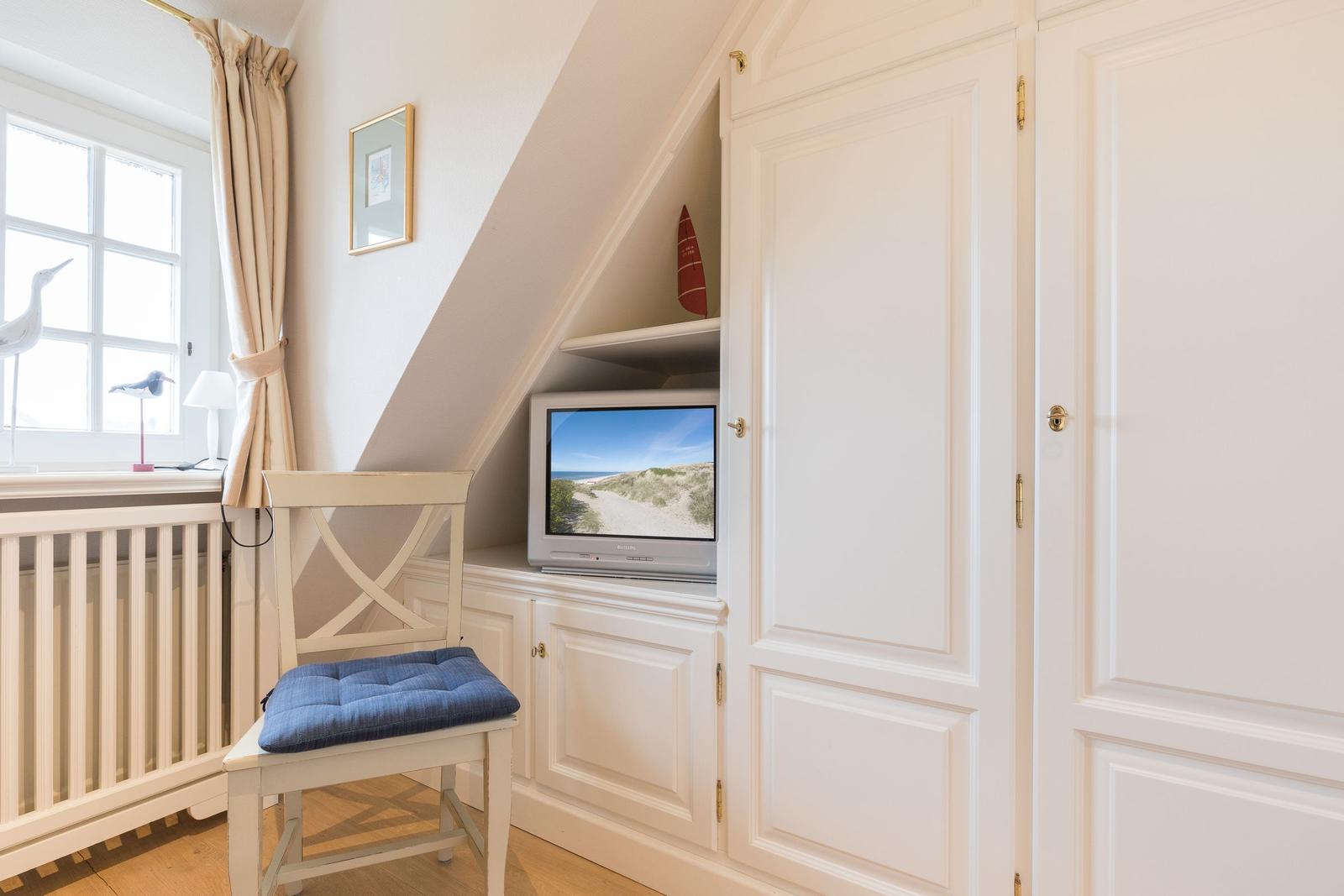 Ein Raum mit weißer Einbauküche, Stuhl mit blauem Sitzkissen und Fernseher unter Dachschräge.