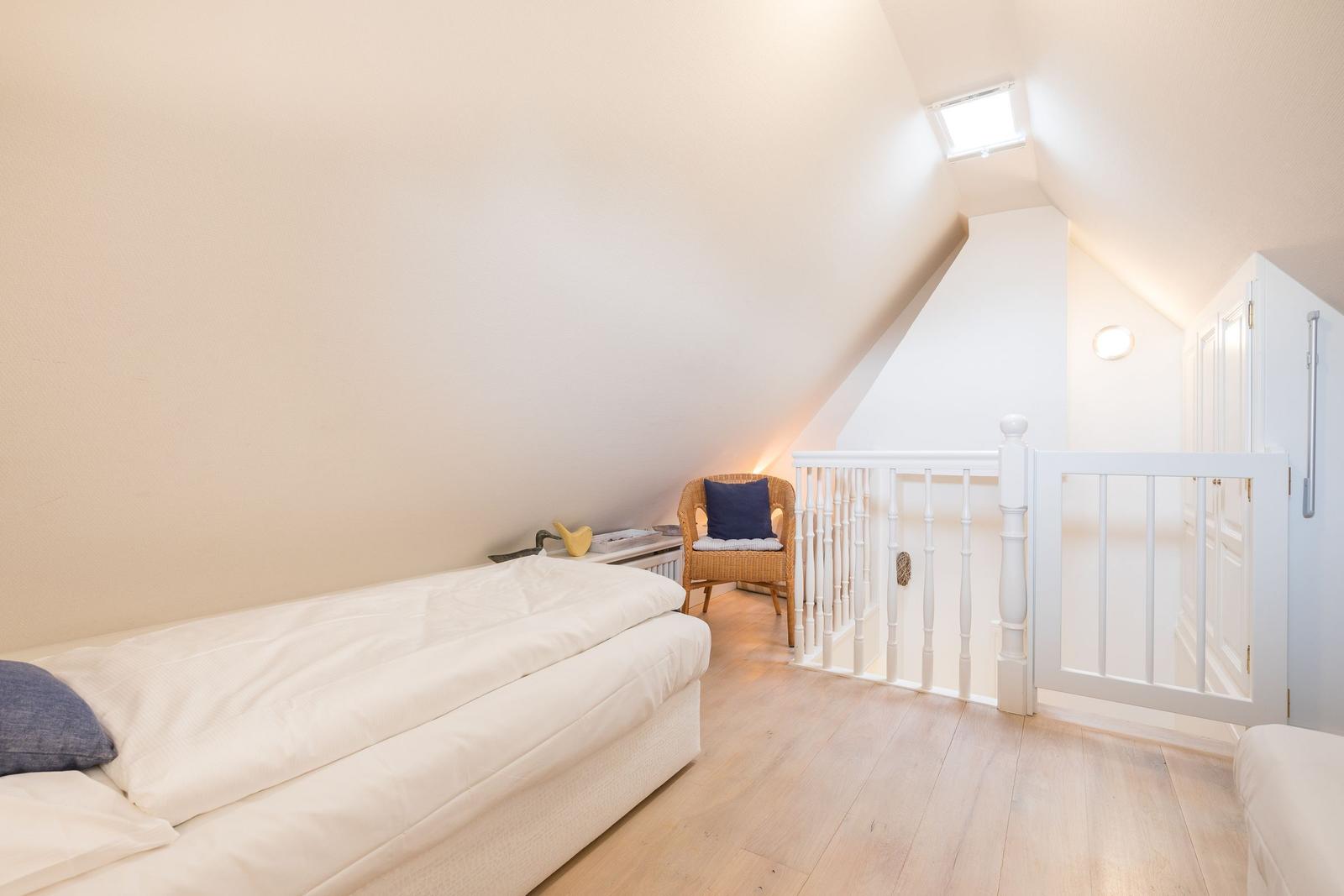 Schlafzimmer mit Dachschrägen, Bett, Stuhl und Sicht auf Treppenhaus.