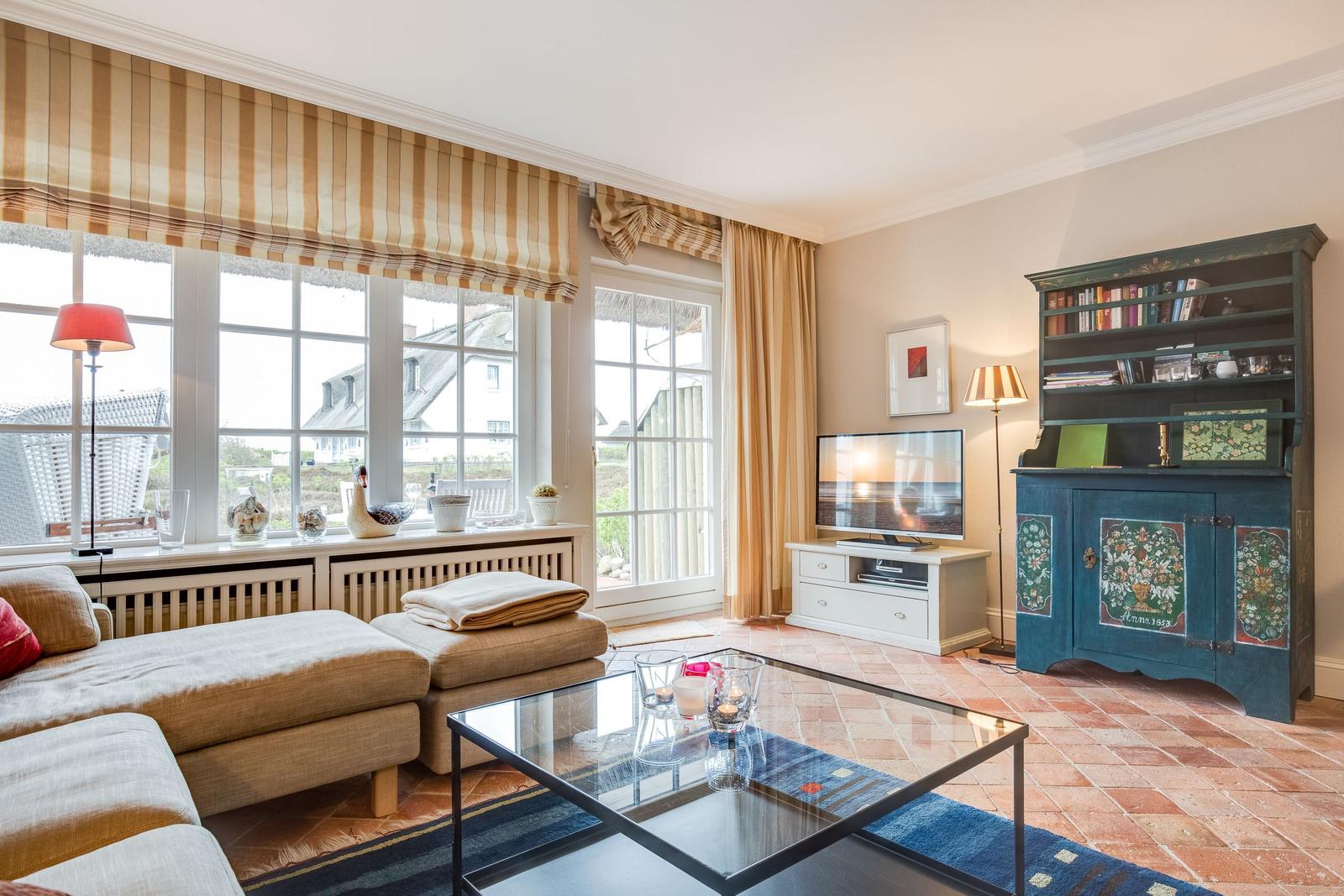 Wohnzimmer mit Sofa, Glas-Tisch und Fernseher. Fenster mit Jalousien und Blick auf Außenbereich.